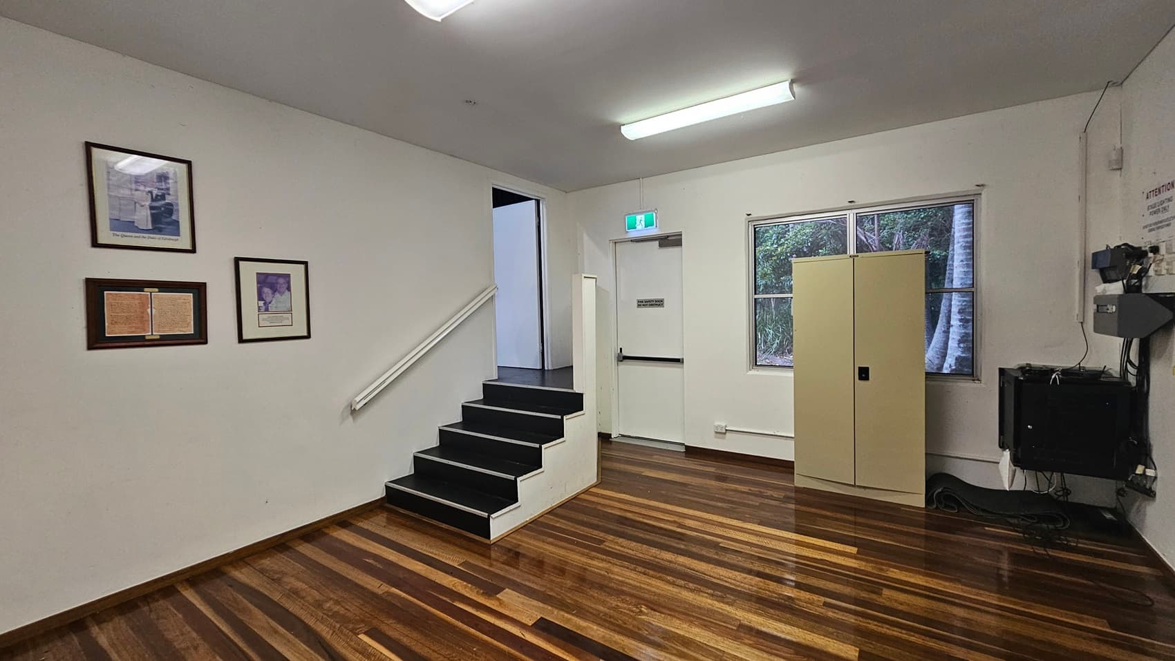 On the left, there are framed pictures on a white wall above a set of black steps with a white handrail. In the middle, a white door with an exit sign above it is slightly open. On the right, a beige cabinet stands next to a window showing greenery outside. The floor is polished wood, and the ceiling has a fluorescent light fixture.