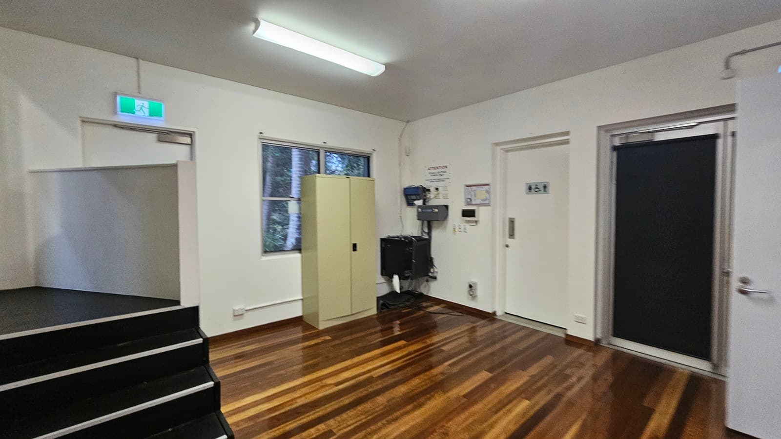 On the left, there are black steps leading to a platform with a green exit sign above a door. In the middle, a beige cabinet stands next to a window showing trees outside. On the right, there are two doors, one with a black curtain and the other with restroom signs. The floor is polished wood, and the ceiling has a fluorescent light fixture.