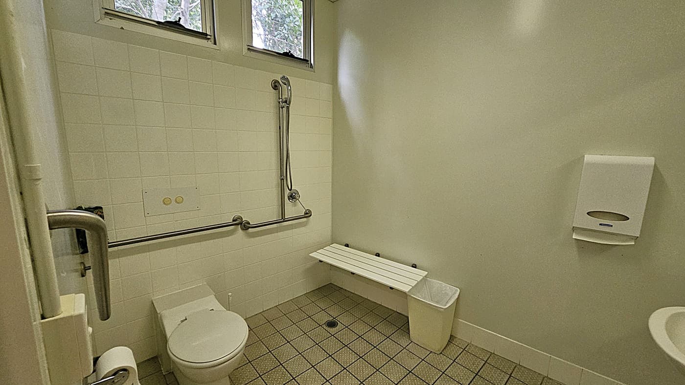 On the left, there is a white toilet with a metal grab rail and a tiled wall featuring a handheld shower. In the middle, a white bench is mounted on the wall above a small waste bin. On the right, a paper towel dispenser is attached to the wall. The floor is covered with small square tiles, and the ceiling is plain with no visible fixtures.