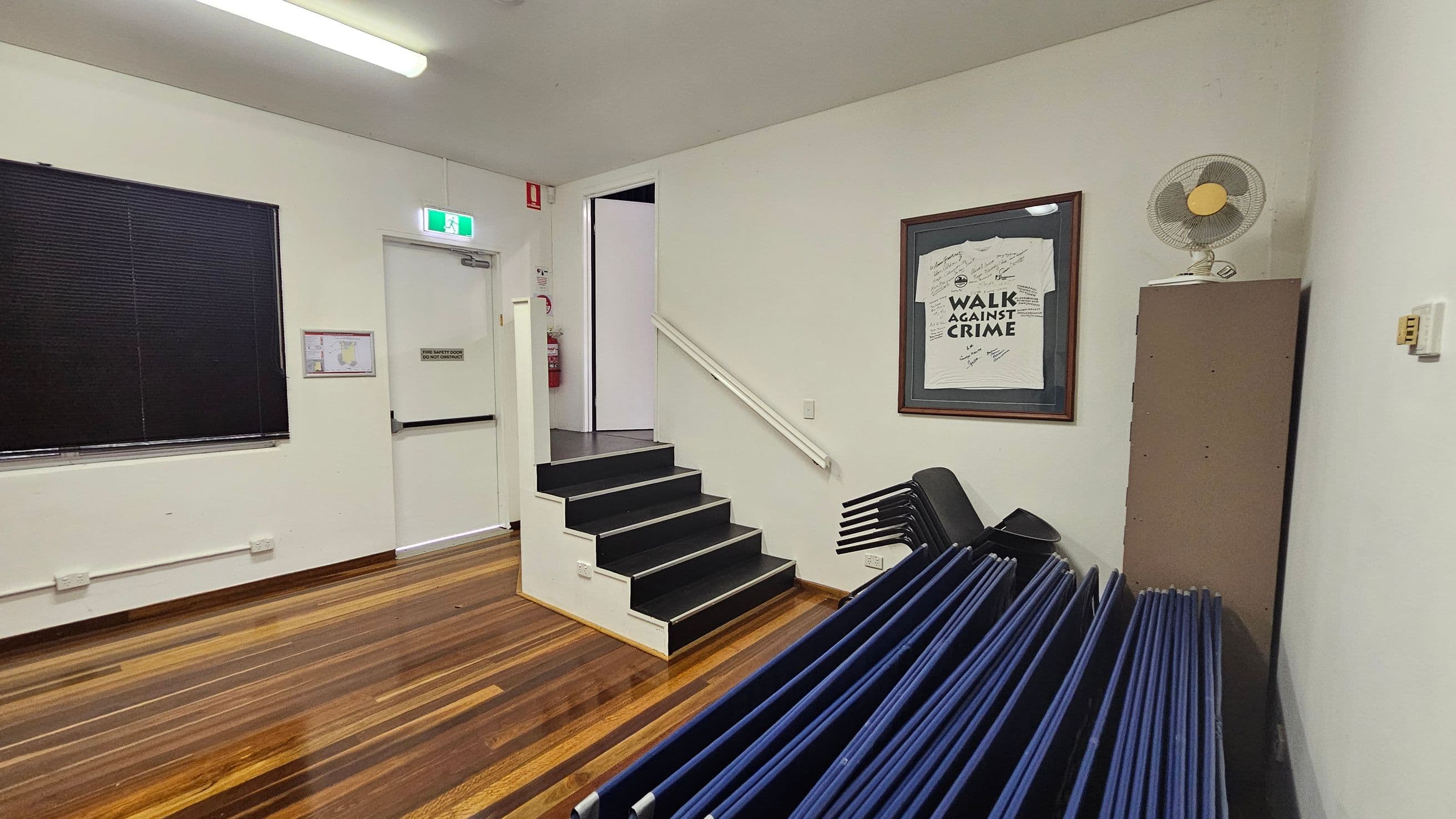 A room with polished wooden floors and a white ceiling. On the left, a window with closed blinds and a door with an exit sign above it. In the centre, a small set of black steps leads to another area. On the right, a framed "Walk Against Crime" shirt is on the wall, with a stack of folded chairs below. A fan sits on top of a tall cabinet. A fire extinguisher is visible near the door.