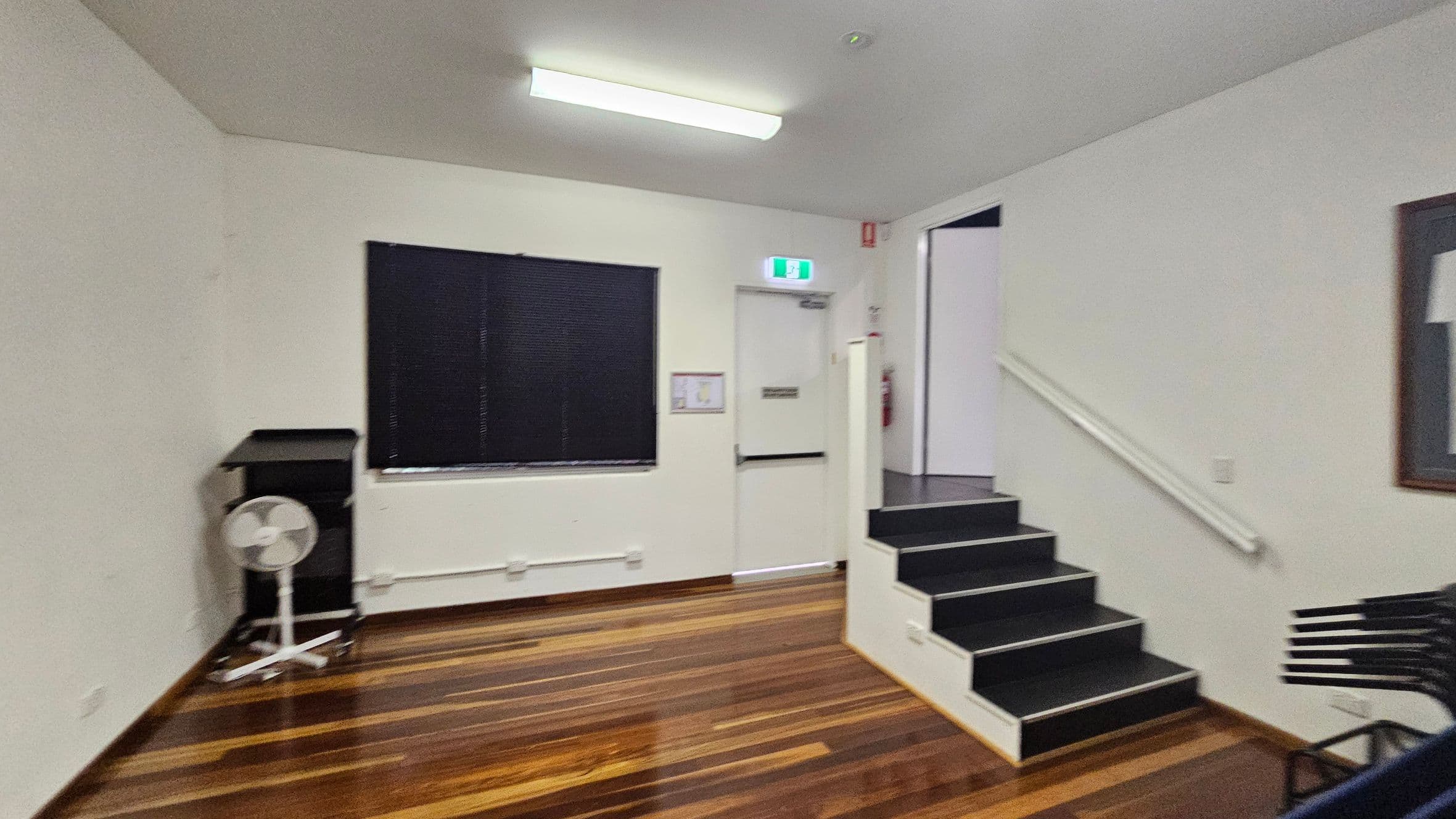 A room with wooden flooring and a white ceiling with a fluorescent light. On the left, there's a black window blind and a fan on a stand. In the centre, a white door with an exit sign above it and a fire extinguisher nearby. On the right, a set of black steps with a handrail leads up to the door. A stack of black chairs is partially visible. A framed picture is on the right wall.