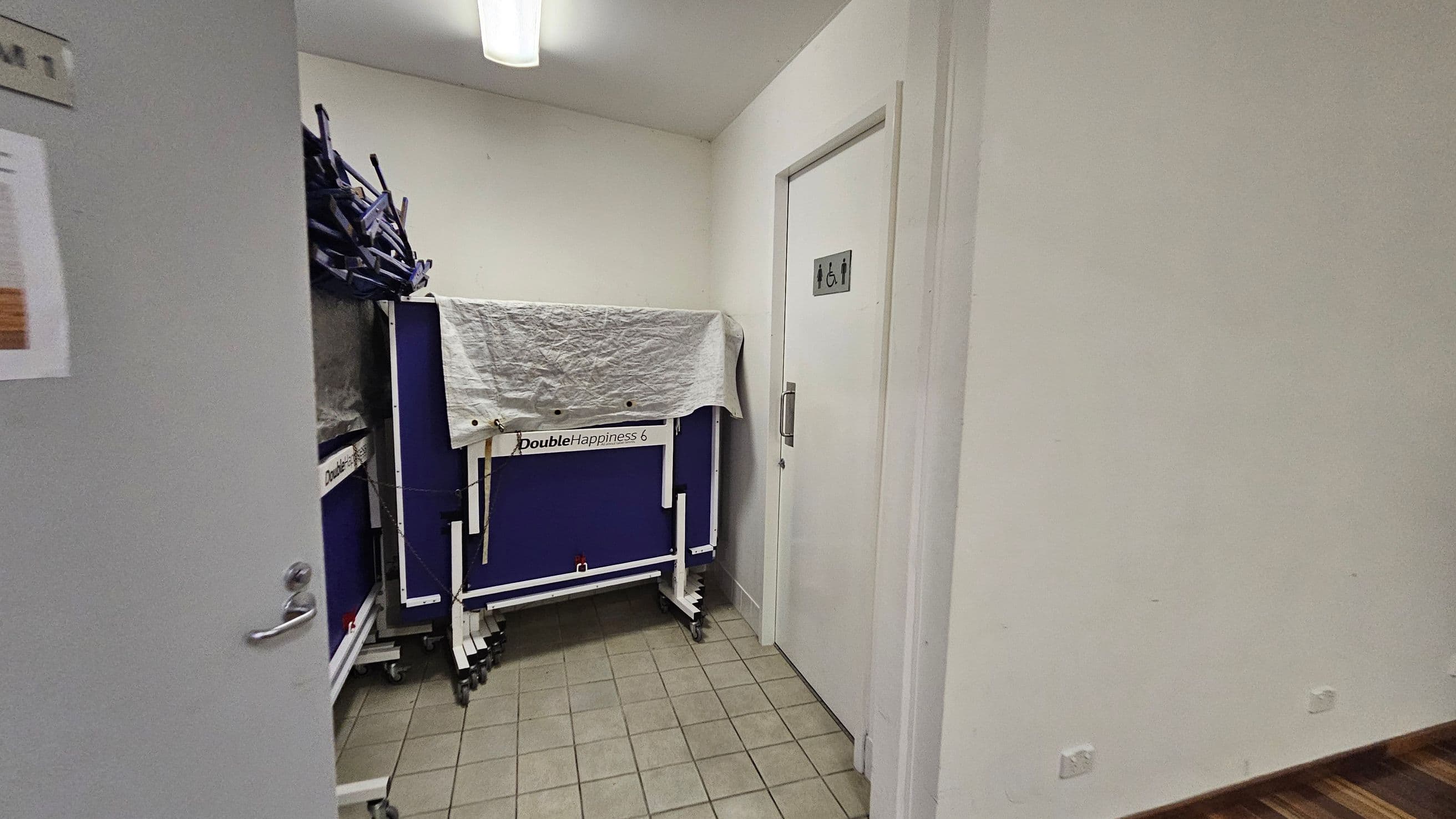 A small room with a tiled floor and a fluorescent light on the ceiling. On the left, there is a partially open door with a sign. In the centre, a folded table tennis table with the label "Double Happiness 6" is covered with a white cloth. On the right, a door with a sign indicating a unisex accessible toilet. The walls are plain and light-coloured.