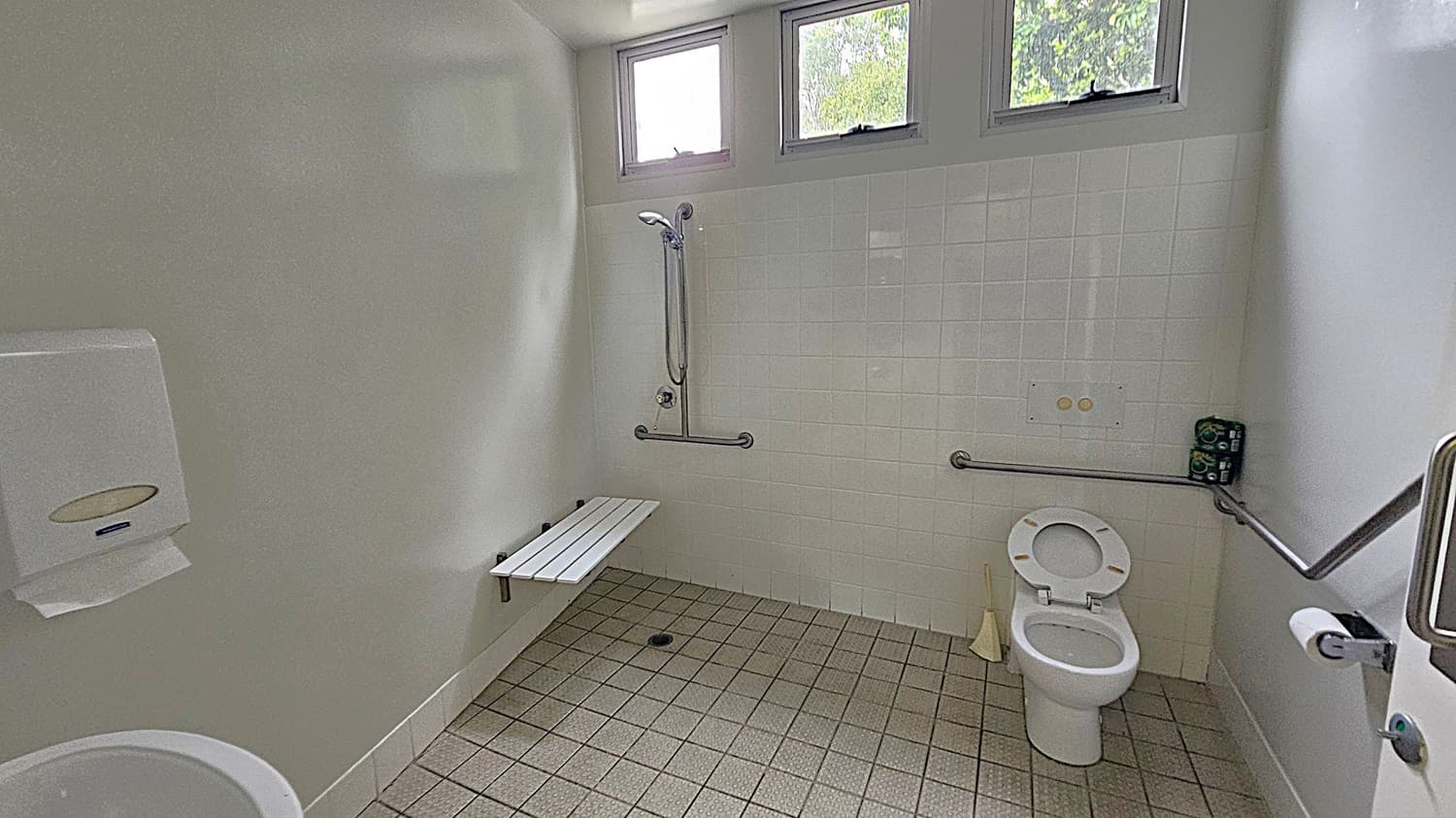 A bathroom with light green walls and a tiled floor. On the left, there's a paper towel dispenser. In the centre, a shower area with a wall-mounted showerhead and a foldable bench. Grab bars are installed around the shower and toilet for accessibility. On the right, a toilet with the lid up, a toilet roll holder, and a small stack of toilet paper. Above, three small windows provide natural light.