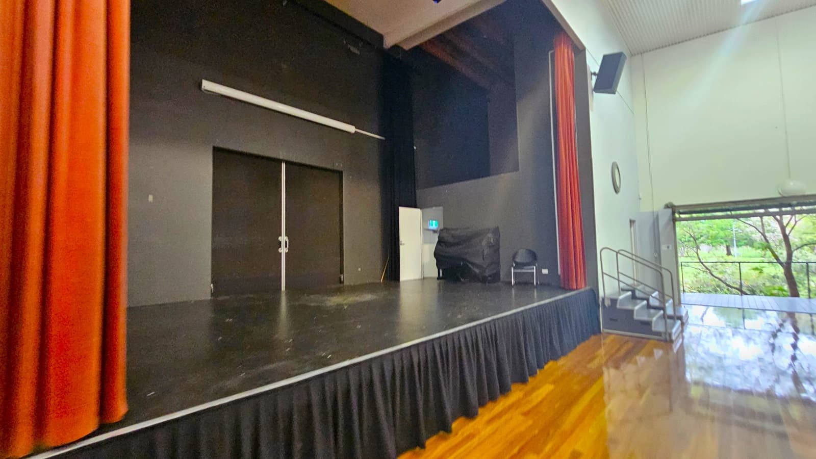 A stage with a black floor and backdrop, flanked by red curtains on the left. On the right, a set of stairs leads down to a polished wooden floor. A chair and a covered object are near the stage's right side. The wall on the right is light-coloured with a clock and a speaker mounted high. Large windows reveal trees outside. The ceiling is high and light-coloured.