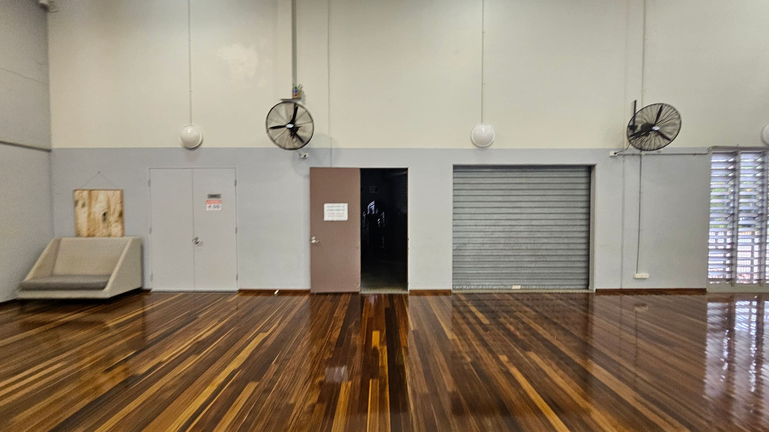 A room with polished wooden flooring and a light green and grey wall. On the left, there's a closed door with a small sign and a cushioned bench. In the centre, an open door leads to a dark space, and a large industrial fan is mounted above. To the right, a closed metal roller door is flanked by another fan. Vertical louvre windows are on the far right, allowing light in. The ceiling is high with hanging spherical lights.