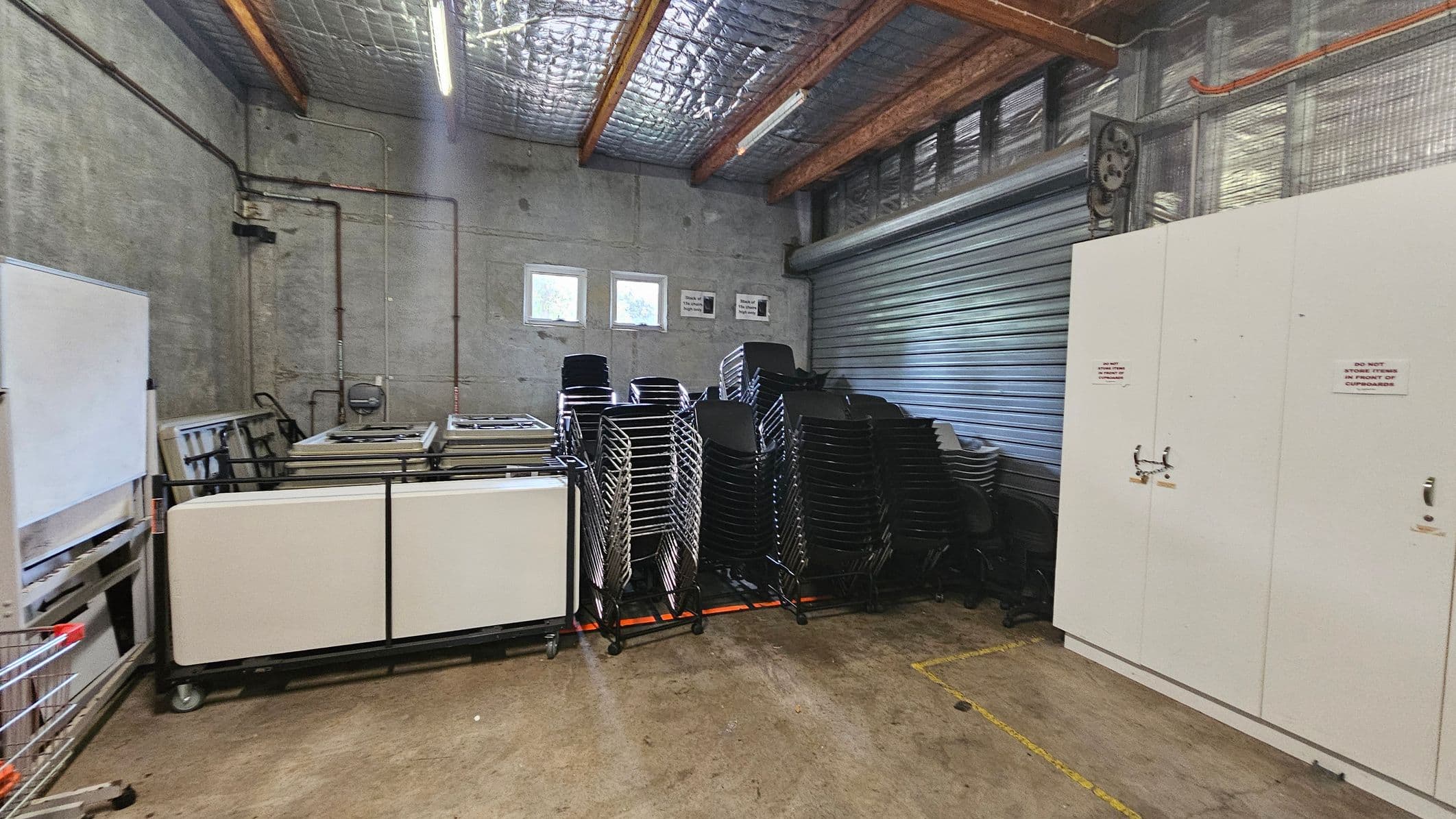 A storage room with a concrete floor and ceiling. On the left, there are stacked tables and a whiteboard. In the centre, numerous black and silver chairs are stacked in rows. On the right, there are large white cupboards with warning signs. The back wall has two small windows and a large metal roller door. The ceiling has exposed beams and insulation.