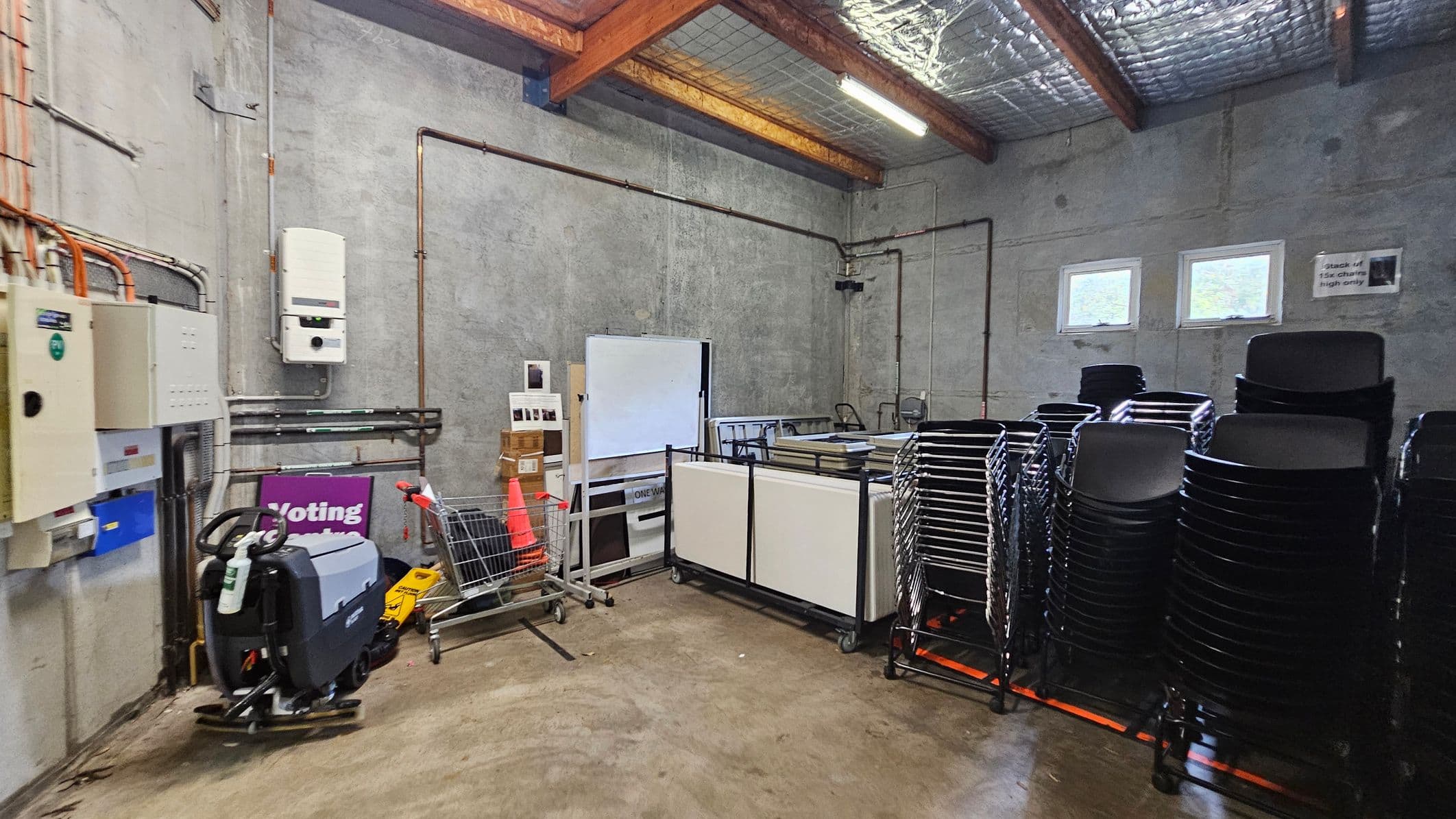 A storage room with concrete walls and a polished concrete floor. On the left, there are electrical panels and a floor cleaning machine. In the centre, a shopping trolley holds a traffic cone and boxes, next to a whiteboard and stacked tables. On the right, there are several stacks of black chairs. The ceiling has exposed wooden beams and insulation, with fluorescent lighting. Two small windows are on the back wall.