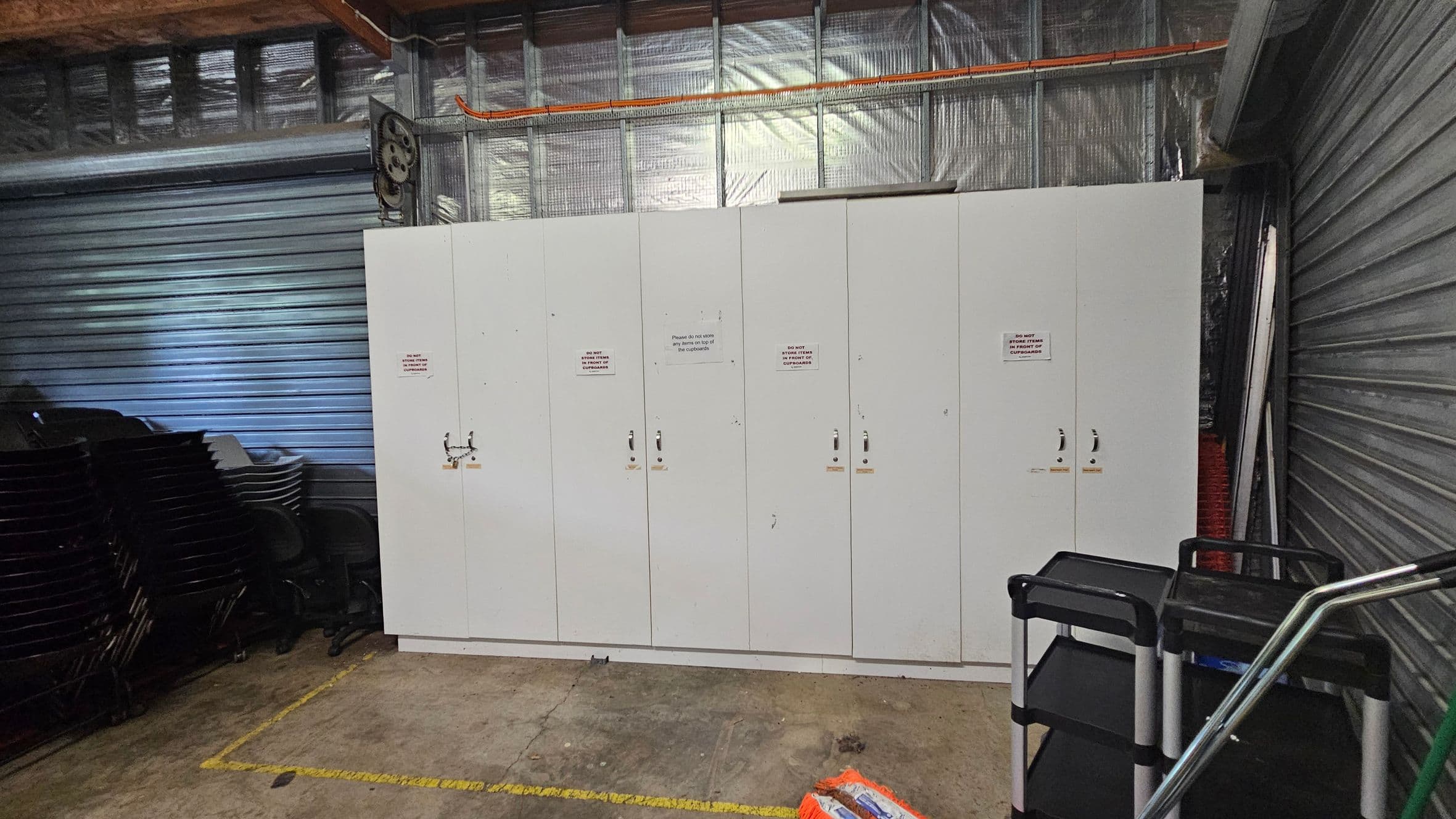 A storage area with a row of white cabinets in the centre, each with a handle and a sign. To the left, there are stacks of black chairs. On the right, there are black trolleys with metal frames. The background features a corrugated metal wall and insulation. The floor is concrete with yellow markings.