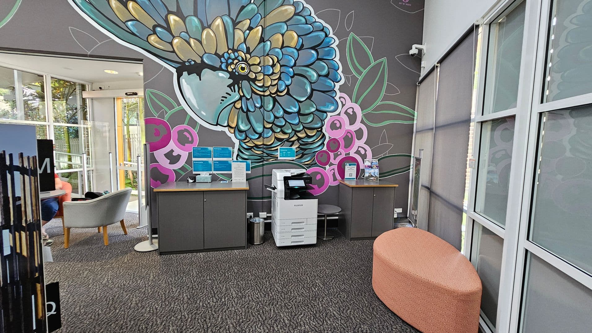A room with a large, colourful mural of a bird on the wall. In the centre, there's a white printer on a grey cabinet, flanked by two more cabinets with informational displays. To the right, a peach-coloured, oval-shaped ottoman sits on a patterned carpet. On the left, a glass door leads to an area with grey chairs and a small table. The ceiling is not visible, and the windows have grey blinds.