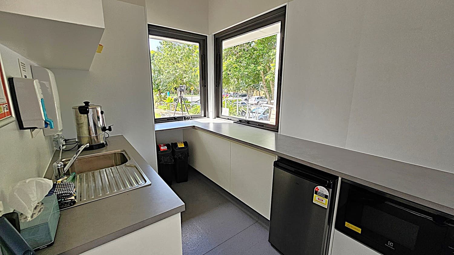 A small kitchen with grey flooring and white walls. On the left, a wooden door is partially open, revealing a wall with control panels labelled "Office", "Meeting Room 2", and "Kitchen". Inside, a countertop runs along the back wall with a small black fridge and a microwave underneath. A large window above the counter provides a view of trees and parked cars outside. A mop is leaning against the right wall.