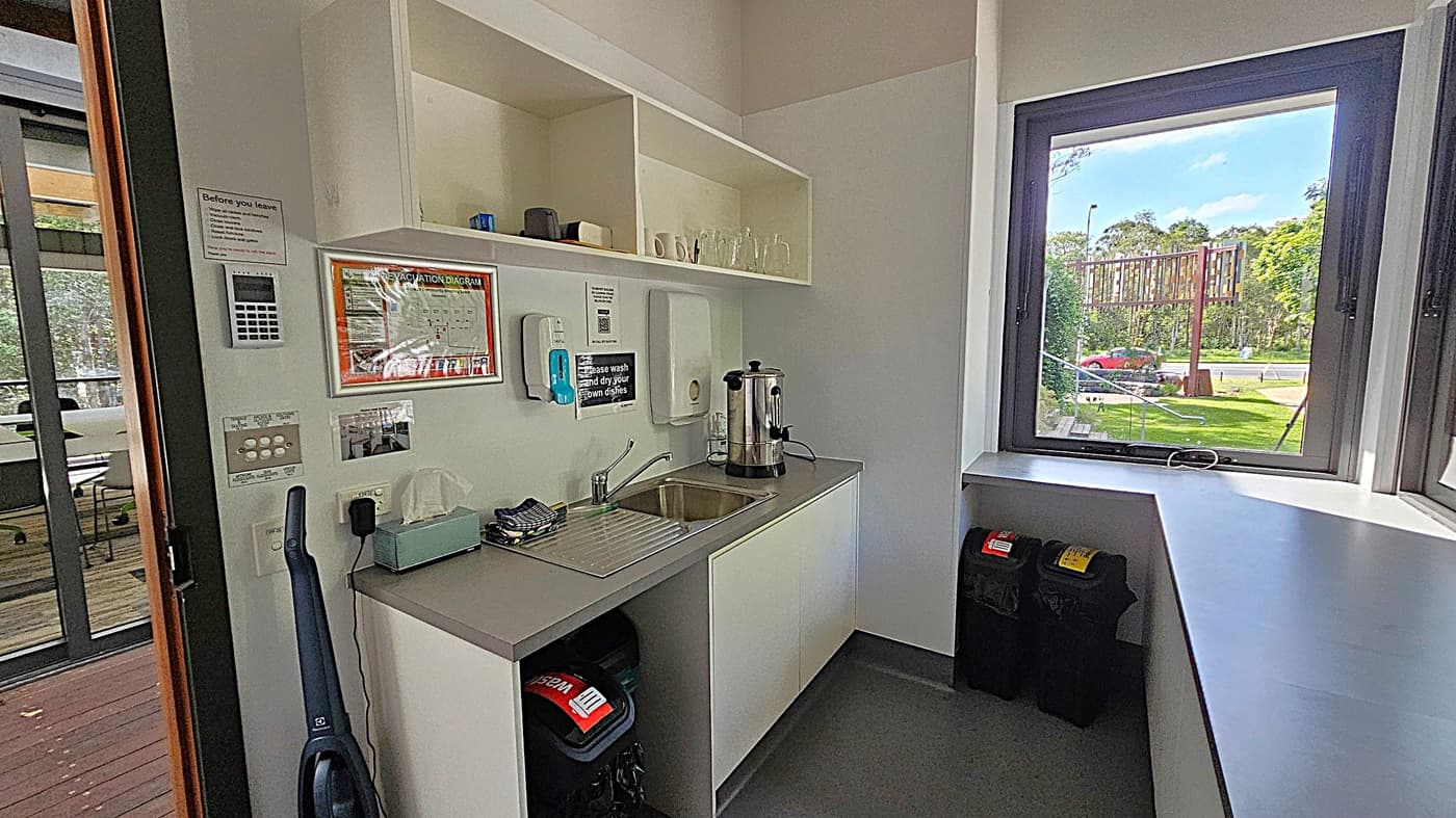 A small kitchenette with a sink and a countertop on the right. Above the sink are open shelves holding mugs and glasses. A large coffee urn is on the counter. On the left wall, there are various notices, a keypad, and a vacuum cleaner. A window on the right offers a view of a grassy area with trees and a road. Below the window, there are two bins. The floor is dark, and the ceiling is plain white.