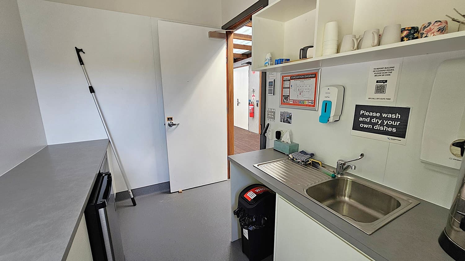 A small kitchen area with a grey countertop and a stainless steel sink on the right. Above the sink, there are shelves holding mugs and other items. A sign reads, "Please wash and dry your own dishes." A mop leans against the left wall. The floor is grey, and the ceiling is plain. An open door leads to a hallway with a visible fire extinguisher and an evacuation diagram on the wall. A waste bin is positioned under the counter.