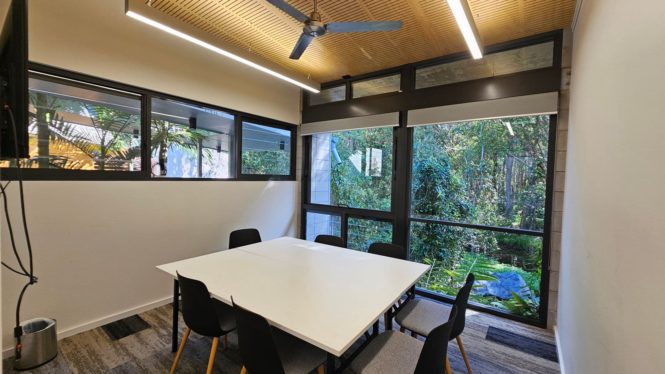 A narrow hallway with a patterned carpet leads to a small meeting room. On the left wall, there's a hand sanitiser dispenser. The room at the end has a white table with black chairs around it. Large windows in the background reveal a view of dense greenery outside. The ceiling is wooden with a linear light fixture and a ceiling fan.