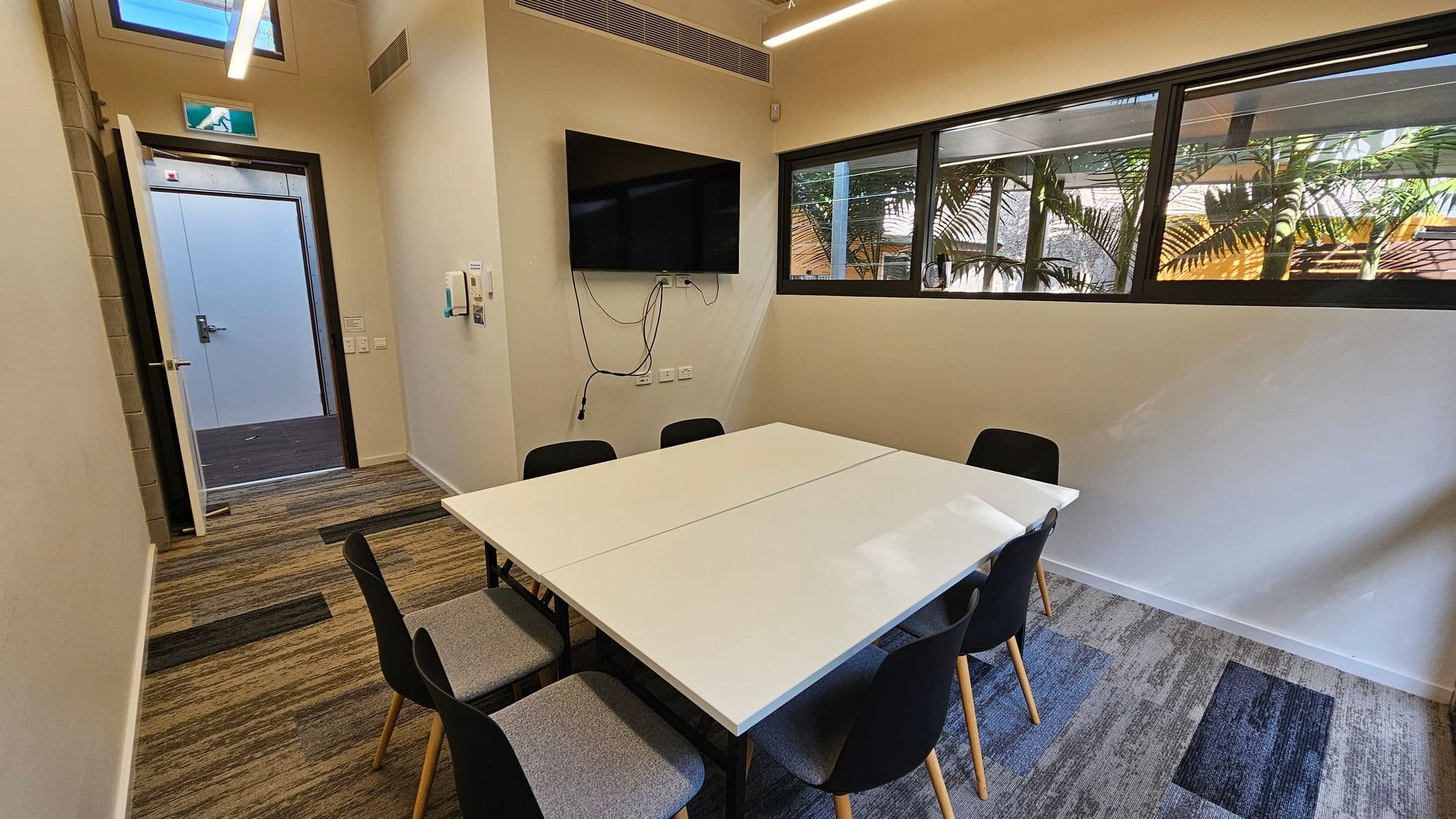 A small meeting room with a rectangular white table surrounded by six black chairs with wooden legs. On the left, an open door leads to a corridor. Above the door, there's an emergency exit sign. A wall-mounted TV with visible cables is on the left wall. The right wall features a row of windows with a view of palm trees outside. The floor is covered with patterned carpet tiles in shades of grey and green. The ceiling has linear lights.