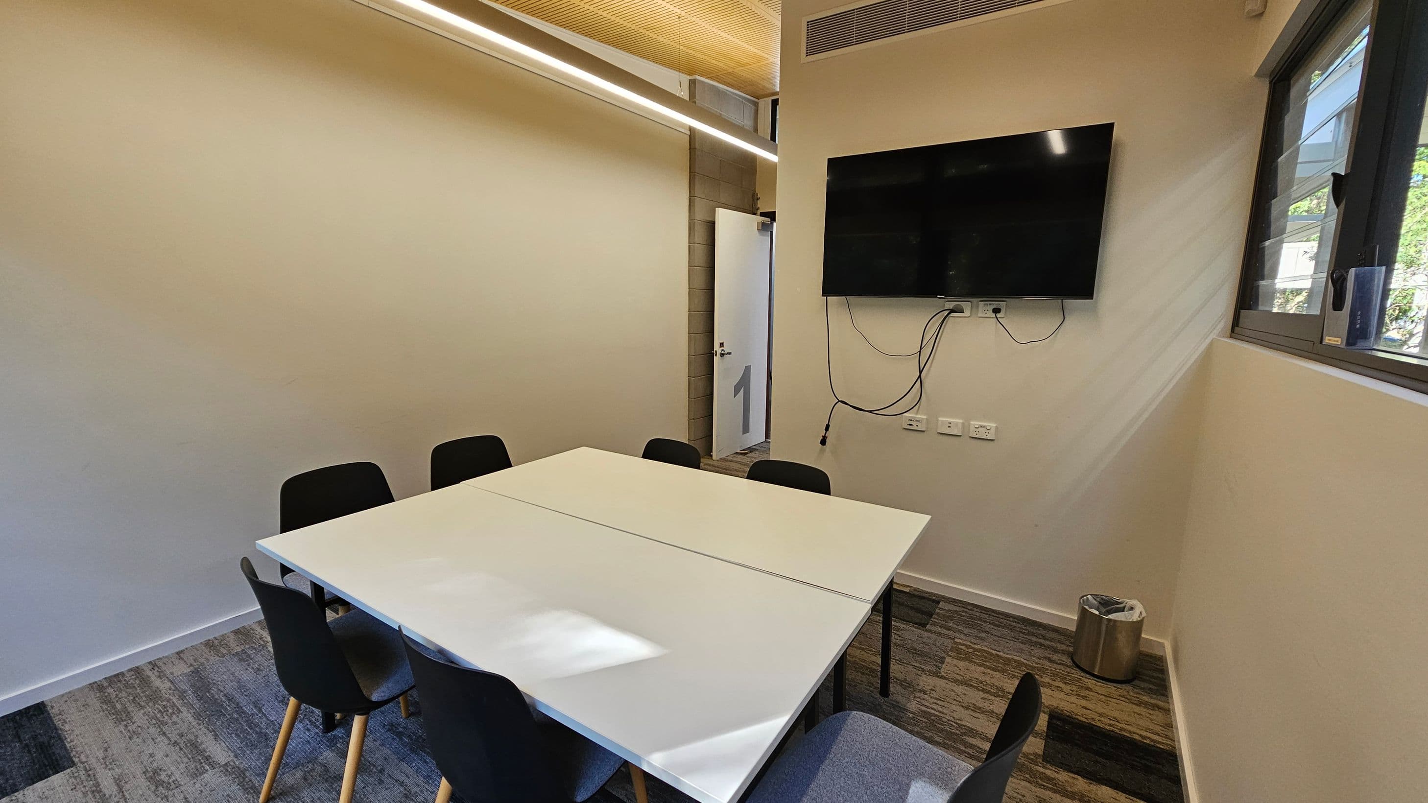 A small meeting room with a rectangular table in the centre surrounded by six black chairs. On the right wall, a flat-screen TV is mounted with visible cables. Below the TV are several power outlets. A window is on the right, allowing natural light in. The floor is carpeted with a mix of grey and green patterns. A metal bin is in the corner. The ceiling has a wooden panel with a strip light. A door is slightly open at the back.
