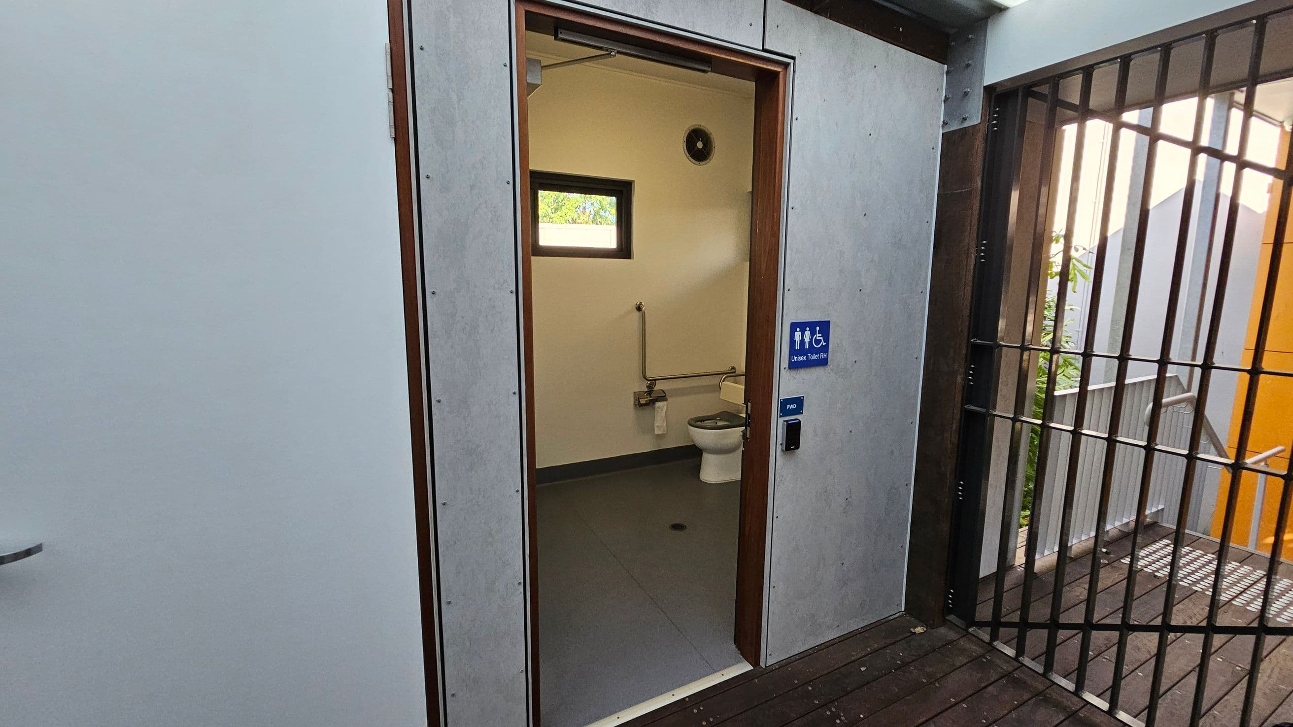 A public toilet with an open door, revealing an accessible restroom inside. The interior has a toilet with grab bars and a small window above. The floor is grey, and the walls are light-coloured. Outside, there is a wooden deck floor and a metal gate on the right. A blue sign on the door indicates it is an accessible toilet.