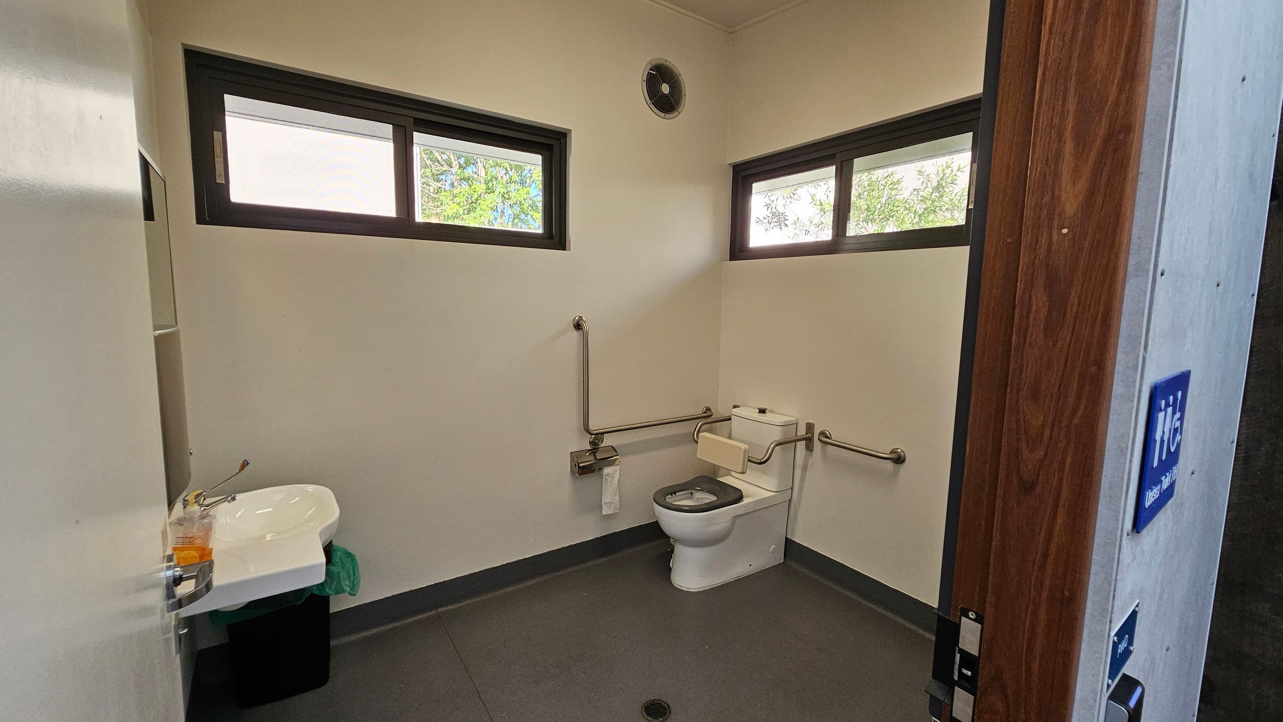 Accessible bathroom with a white toilet in the corner, equipped with metal grab bars on both sides. A small sink with a tap is on the left wall, above a black bin. The walls are light-coloured, and there are two rectangular windows near the ceiling, allowing natural light in. A ventilation fan is above the windows. The floor is grey, and the door is wooden with a blue accessibility sign visible.