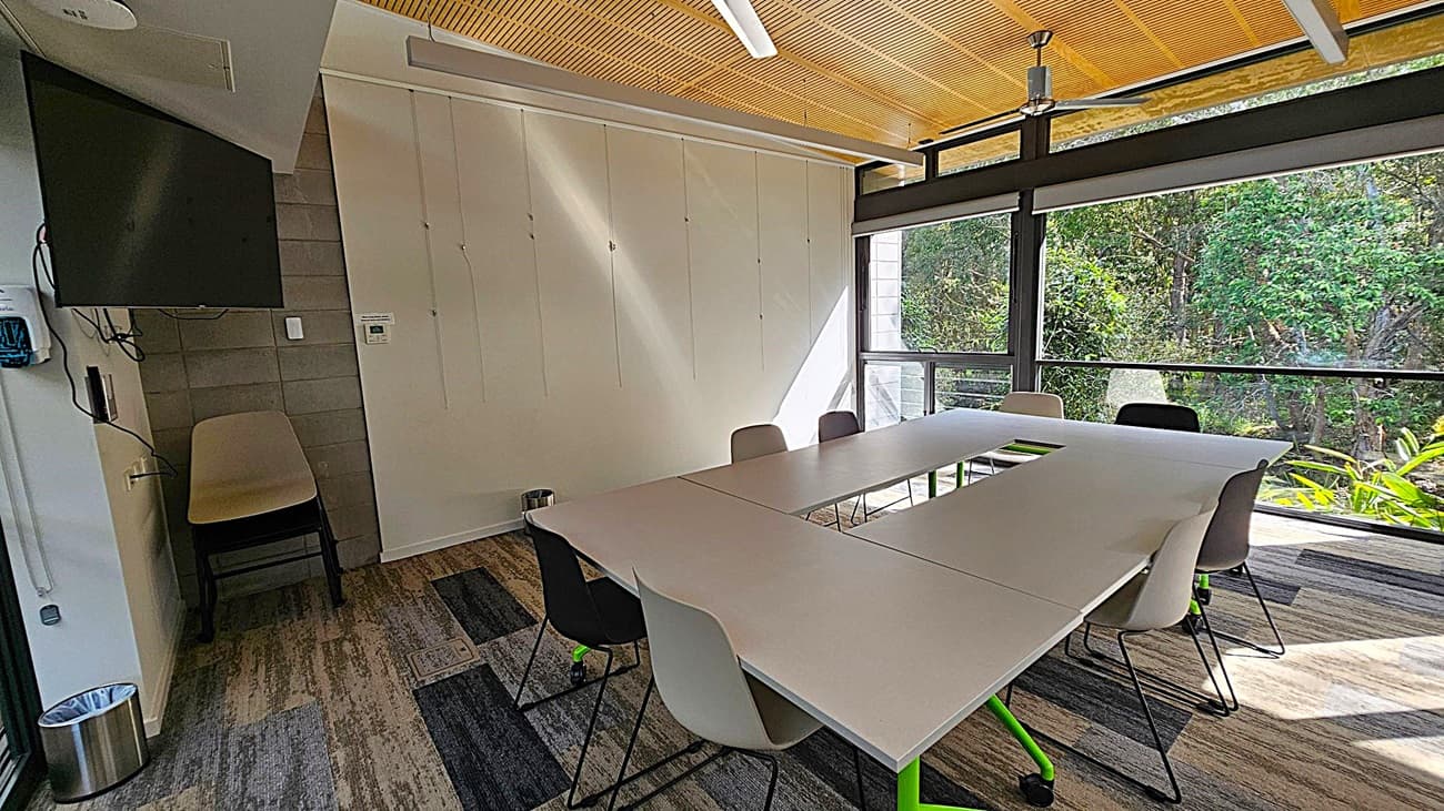 A modern meeting room with a U-shaped table arrangement and several chairs. On the left, a wall-mounted TV and a stack of chairs are visible. The floor has a patterned carpet in shades of grey and green. The ceiling is wooden with a light fixture. Large windows on the right provide a view of lush greenery outside, allowing natural light to fill the room. A small waste bin is placed near the wall.