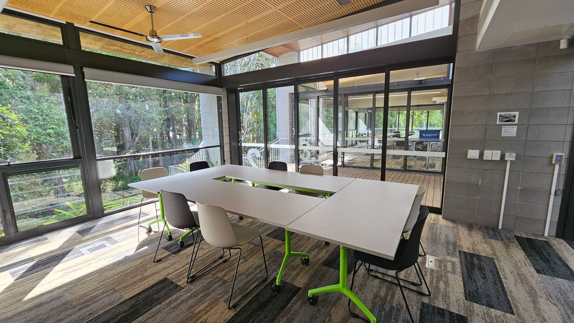 A modern meeting room with large glass windows and doors, offering a view of lush greenery outside. In the centre, there is a U-shaped table with white surfaces and green legs, surrounded by a mix of black and beige chairs. The floor is covered with a patterned carpet in shades of grey and brown. The ceiling features wooden panels and a ceiling fan. On the right, a grey brick wall has some notices and switches.