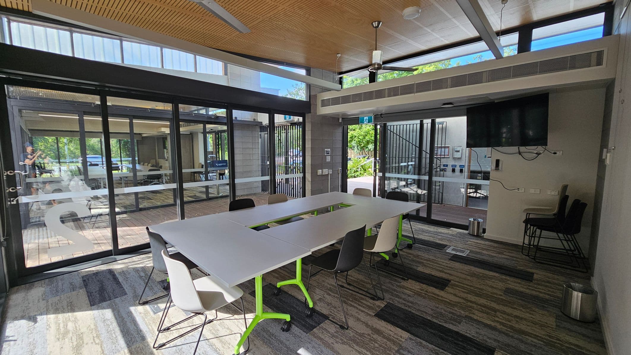 A modern meeting room with a U-shaped table in the centre, surrounded by chairs. The floor is covered with patterned carpet tiles. On the right, a wall-mounted TV with visible cables and stacked chairs. Large glass doors on the left and background open to an outdoor area with greenery. The ceiling is wooden with a fan and recessed lighting.