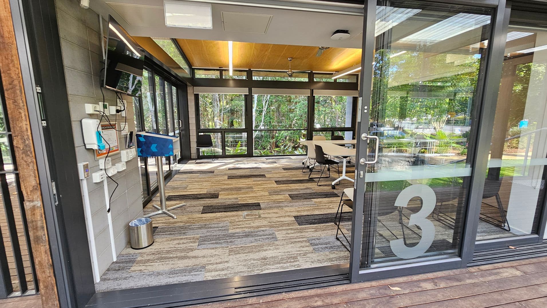 A modern meeting room with large glass sliding doors, revealing a long table surrounded by chairs on a patterned carpet. The room is well-lit with natural light from the surrounding windows. Outside, there's a wooden deck with a transparent roof and a seating area with benches. The area is surrounded by lush greenery, visible through the glass. The number "3" is marked on the glass door.