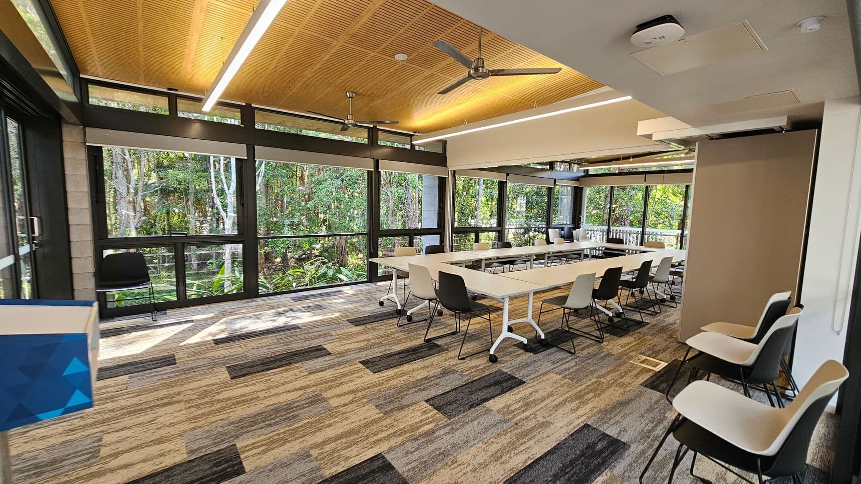 A modern conference room with large windows on the left and right, offering views of lush greenery outside. The room features a long, rectangular table with white surfaces and a mix of black and white chairs. The floor is covered with a patterned carpet in shades of grey, beige, and green. The ceiling is wooden with recessed lighting and ceiling fans. A blue and white object is partially visible in the foreground on the left.
