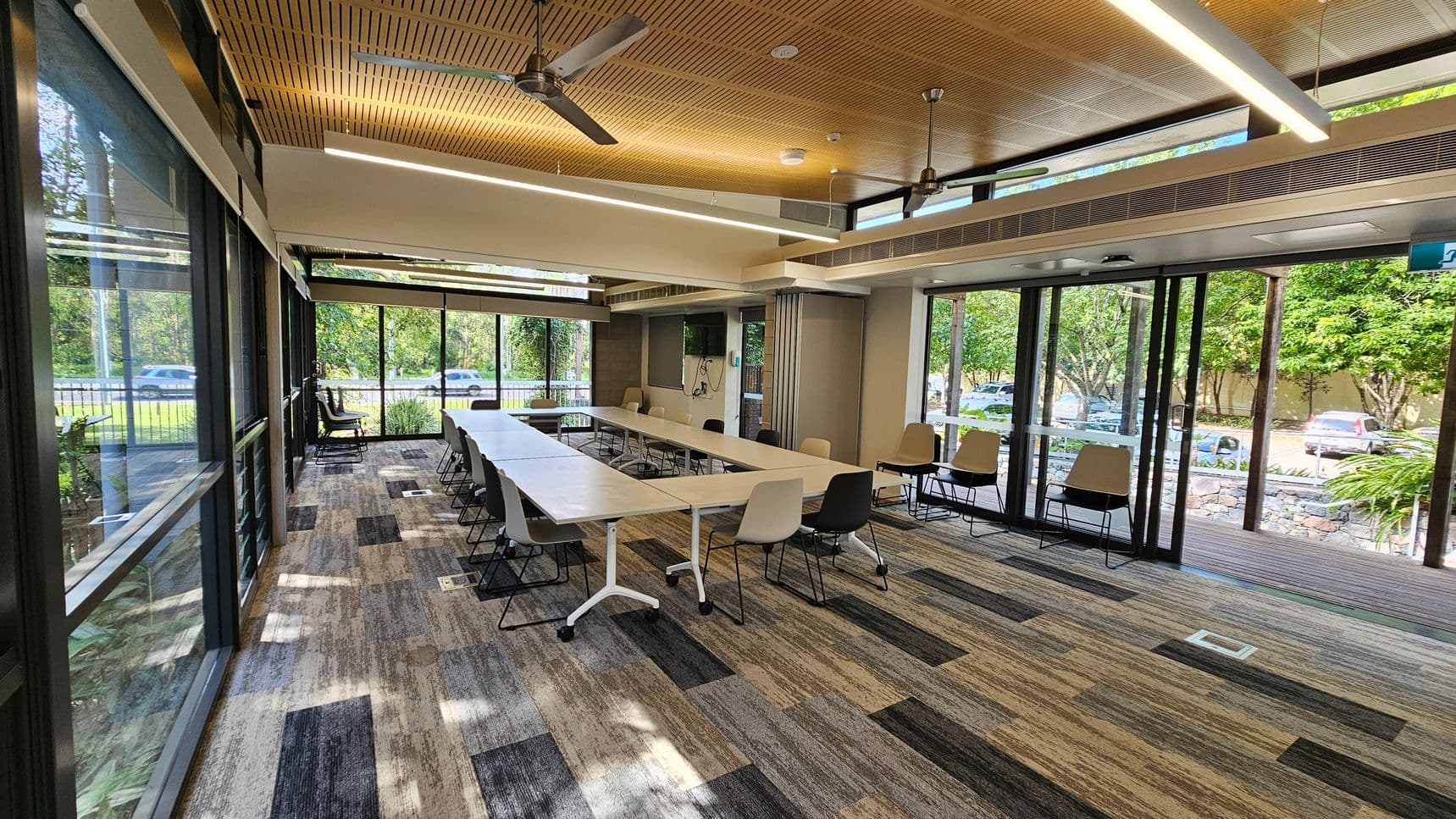 A modern conference room with large windows on the left and right, allowing natural light to flood in. The room features a long, white, modular table with various chairs around it. The floor is covered with a patterned carpet in shades of grey and beige. The ceiling is wooden with recessed lighting and ceiling fans. Outside, there is a view of greenery and parked cars. The room has sliding glass doors leading to an outdoor area with more greenery visible.
