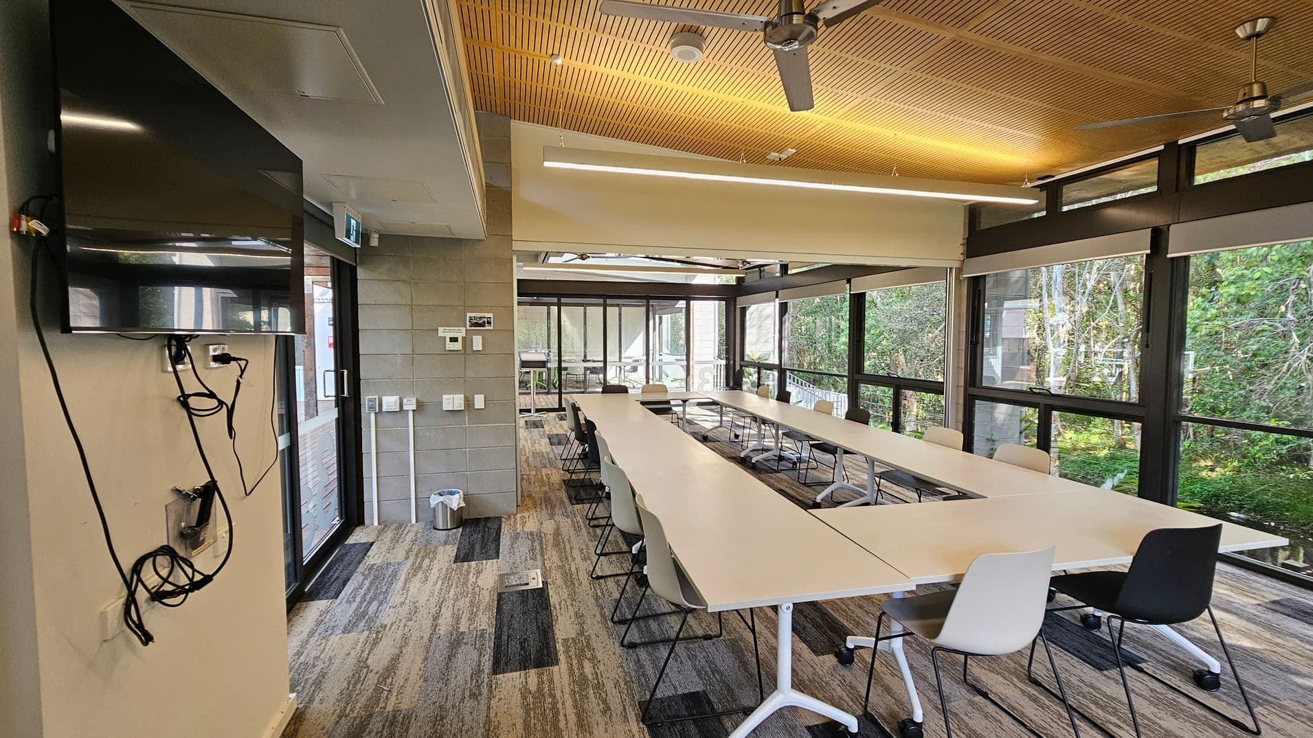 A modern conference room with a U-shaped arrangement of tables and chairs. On the left, a large TV is mounted on the wall with visible cables. The room has large windows on the right, offering a view of greenery outside. The ceiling is wooden with ceiling fans and strip lighting. The floor is carpeted with a patterned design. At the far end, there are glass doors leading to an outdoor area.