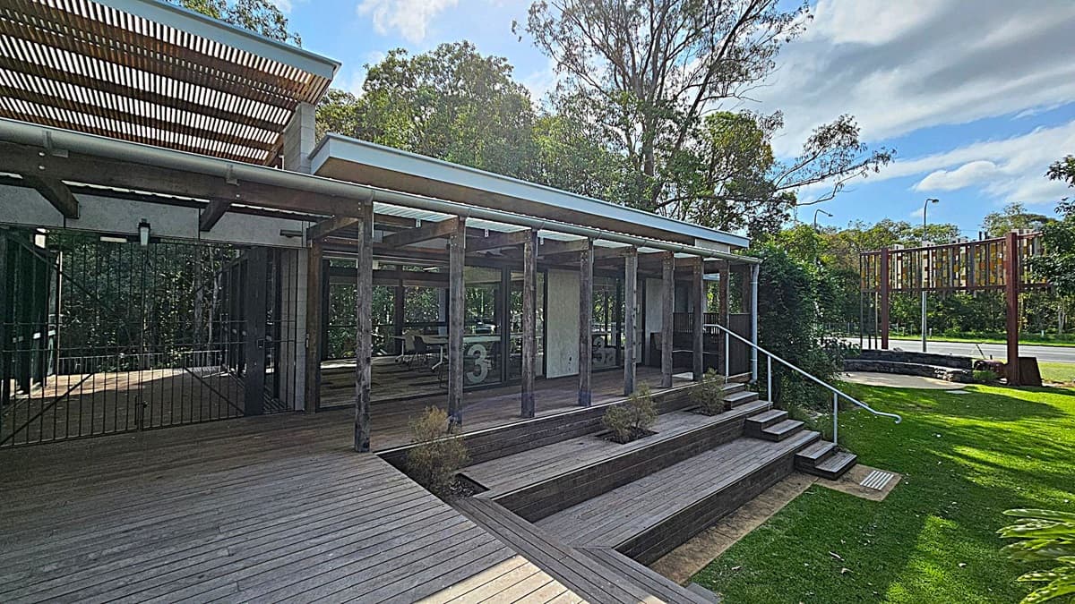A modern building with corrugated metal walls and a wooden decked floor. On the left, there's a small bench and a window with a colourful mural visible inside. The entrance is in the centre, featuring a door with a sign and a fire extinguisher symbol. Above, a slatted wooden awning provides partial shade. To the right, an open area with tables and chairs is visible, surrounded by trees in the background.