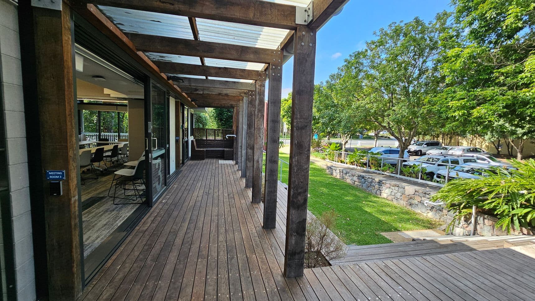 A wooden deck extends along the left side, with a meeting room visible through large glass doors. The deck is covered by a transparent roof supported by wooden beams. On the right, a grassy area with a stone wall and lush green plants leads to a row of parked cars. Trees line the background, providing shade and greenery.