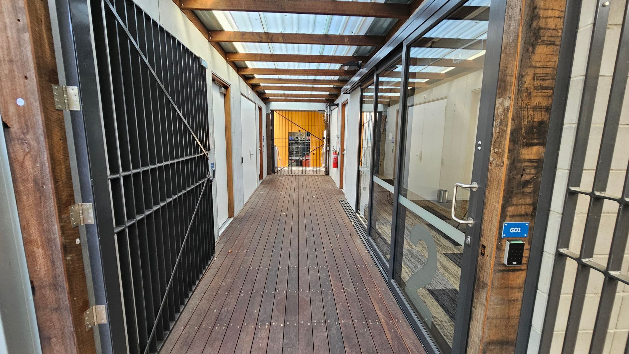 A narrow corridor with a wooden floor and a transparent roof supported by wooden beams. On the left, there is a large metal gate and a white door. On the right, a glass wall with a door marked "G01" and a keypad entry system. At the end of the corridor, a metal gate leads to a space with a yellow wall and visible shelving. A fire extinguisher is mounted on the right side near the end.