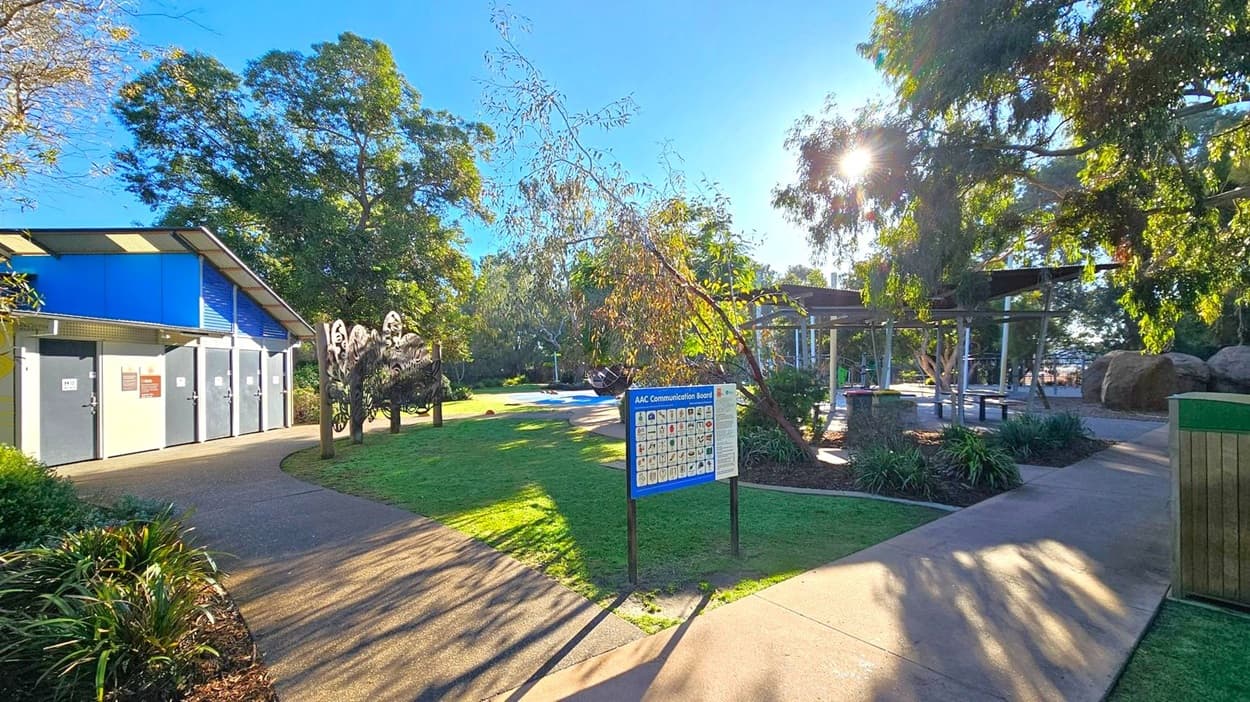 A park scene with a building on the left featuring grey doors and a blue roof. In the centre, there's a sign labelled "AAC Communication Board" on a grassy area. To the right, a shaded area with tables and benches is partially covered by a metal structure. Trees surround the area, with sunlight filtering through the branches, casting shadows on the ground. The path is paved, and there are some plants and bushes along the edges.