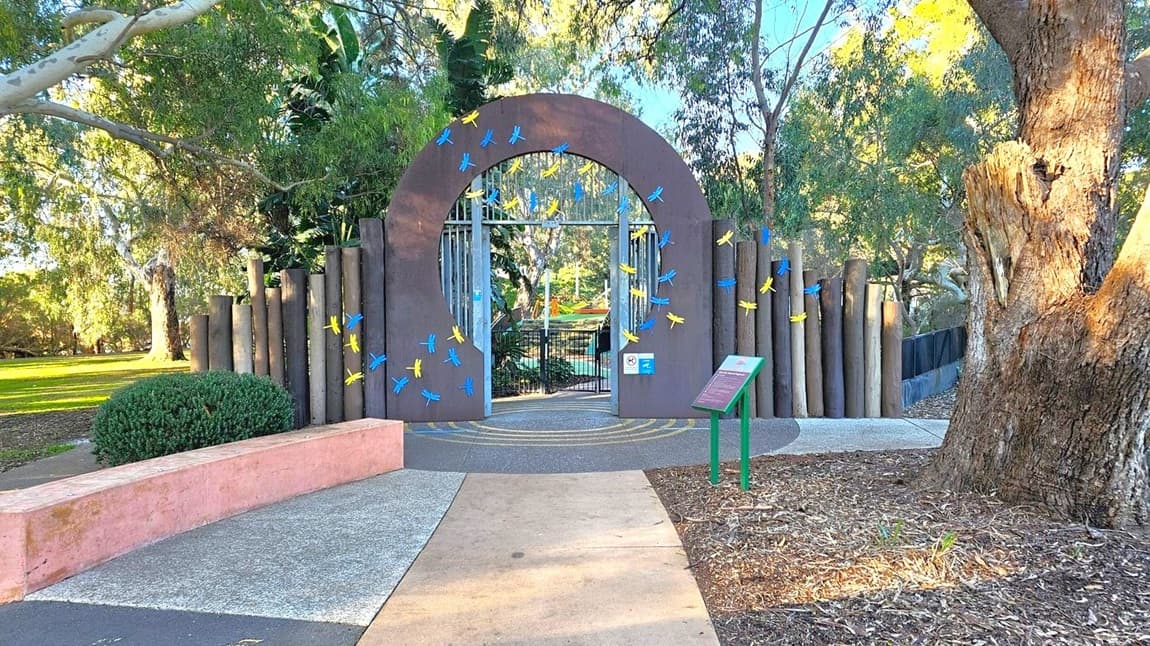 A large, circular archway decorated with blue and yellow dragonfly motifs stands in the centre, leading to a gated entrance. The arch is flanked by vertical wooden posts. On the left, there's a curved pink concrete bench and a bush. On the right, a large tree trunk and a green signpost with a slanted information board. The ground is a mix of concrete and mulch, with trees and greenery in the background.