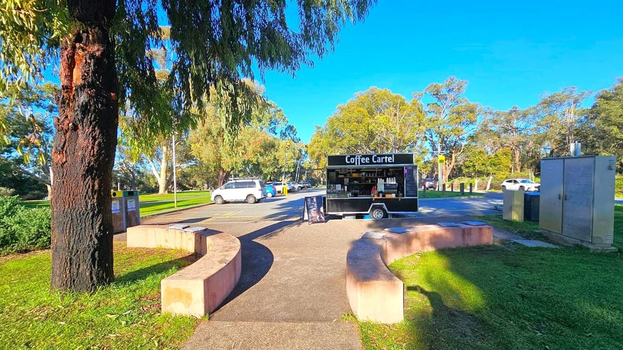 A coffee cart labelled "Coffee Cartel" is positioned in a car park area. In the foreground, there's a curved stone bench and a large tree with a textured trunk. To the left, there are rubbish bins near the tree. On the right, a utility box is visible. Several cars are parked in the background, with trees and greenery surrounding the area. The ground is a mix of grass and pavement.