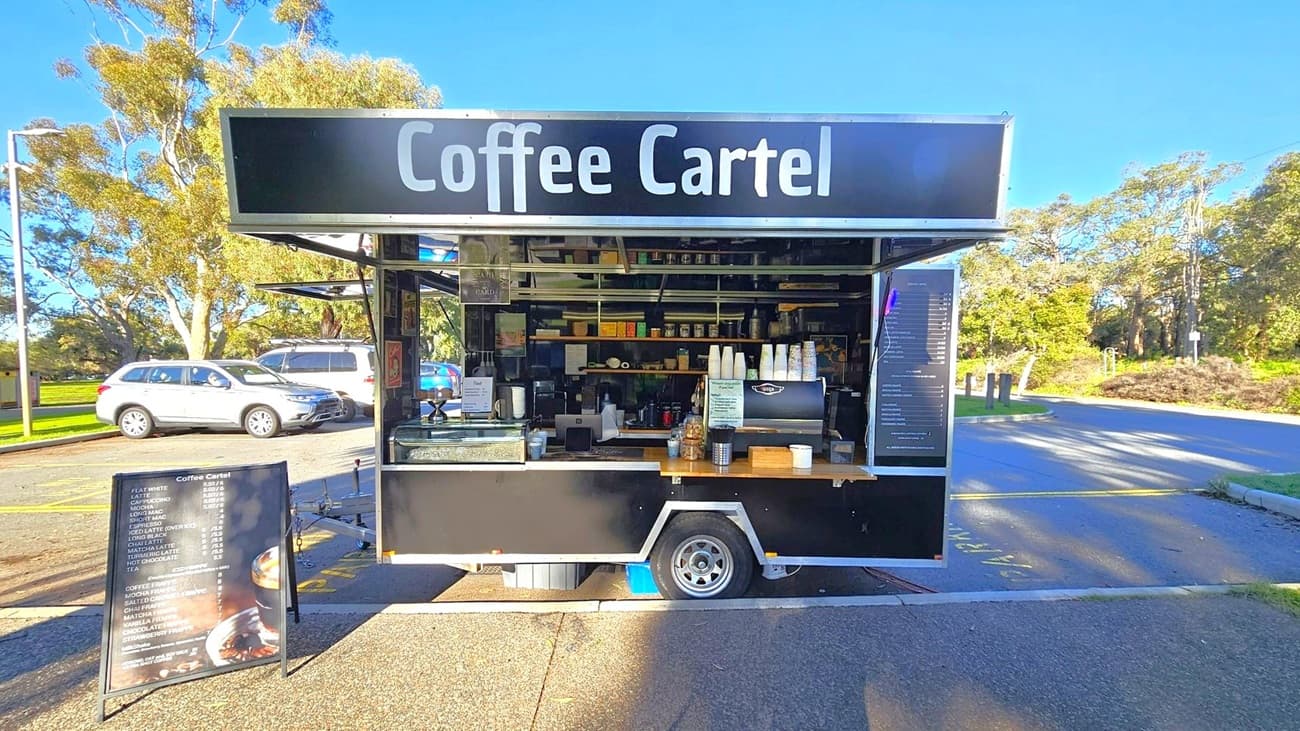 A black coffee cart with the sign "Coffee Cartel" at the top. The cart has an open serving area displaying coffee cups, a coffee machine, and various supplies. A menu board is placed on the ground in front of the cart. To the left, there are parked cars and trees, while the right side shows a road and more greenery. The ground is paved, and the sky is clear.