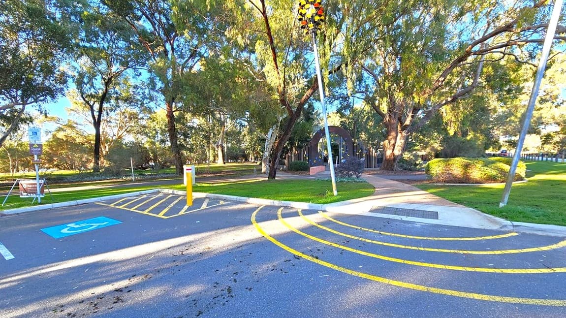 A parking area with a designated disabled parking space marked in blue on the left. Yellow lines curve across the asphalt. A path leads from the parking area into a park with lush green grass and tall trees. A pole with a cluster of orange and black objects is on the right. In the background, there is a structure with an arched entrance surrounded by trees and bushes. A signpost is visible near the parking space.