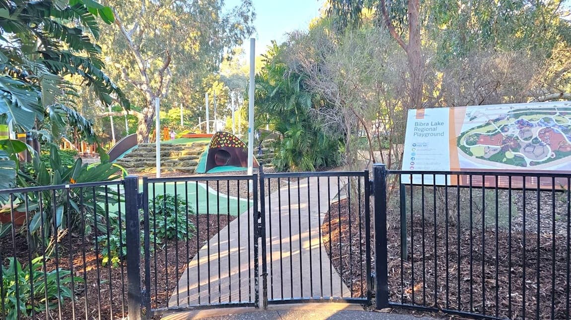 A black metal gate in the foreground opens to a path leading into a playground. On the right, a large sign displays a map of Bibra Lake Regional Playground. The area is surrounded by lush greenery, including tall trees and various plants. In the middle, a small tunnel structure and climbing steps are visible. The ground is covered with mulch and the path is paved.