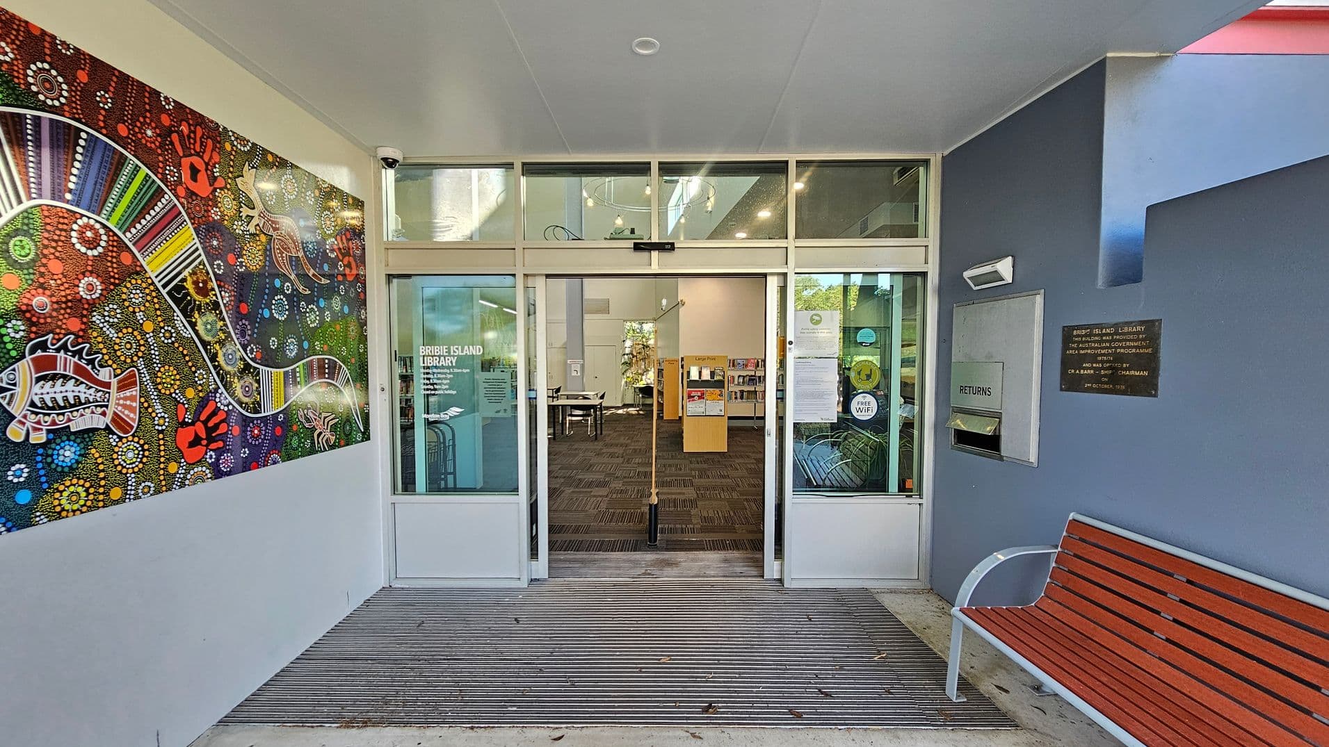 A library entrance with glass doors in the centre, labelled "Bribie Island Library". To the left, a vibrant mural features colourful Aboriginal art with patterns and animal motifs. On the right, a blue wall has a book return slot and a plaque. A wooden bench with metal armrests is positioned below. The floor is a combination of concrete and a ribbed mat leading into the library. The ceiling is plain with a light fixture.