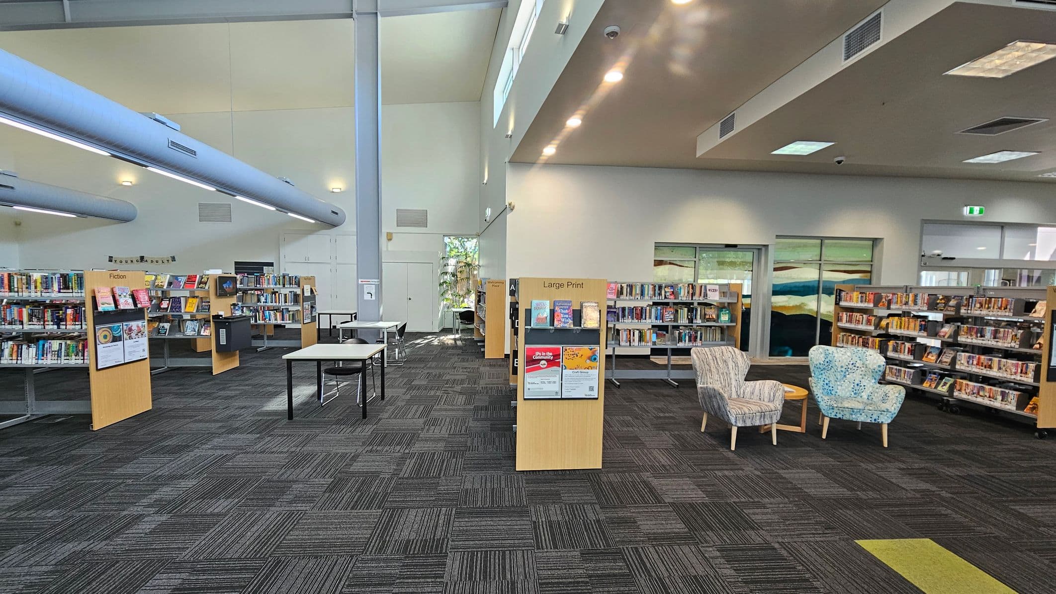 A library interior with a grey carpeted floor. On the left, there are bookshelves labelled "Fiction" filled with books. In the centre, a section labelled "Large Print" is visible, along with a couple of tables and chairs. On the right, there are more bookshelves and two patterned armchairs with a small round table between them. The ceiling has a mix of recessed and fluorescent lighting, and large windows allow natural light in.