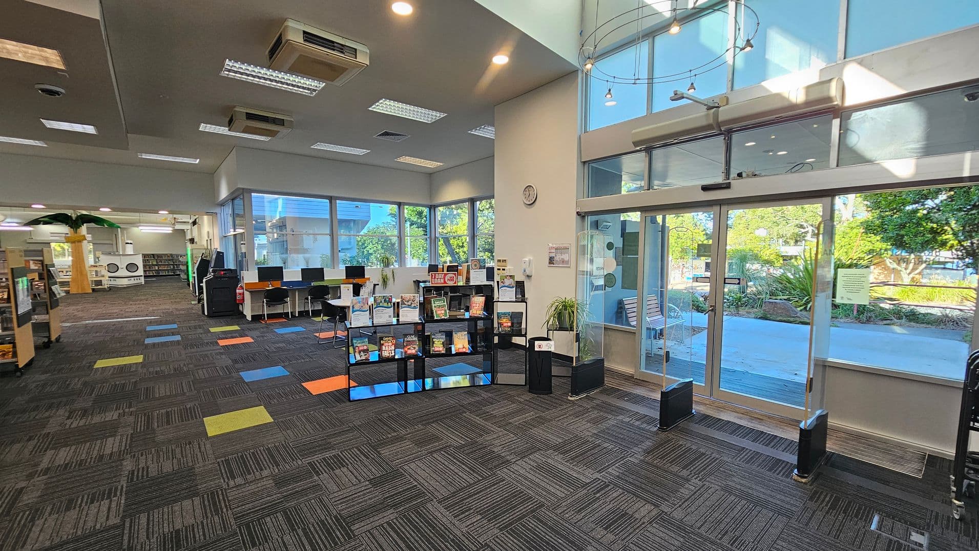 A library interior with a patterned carpet featuring colourful squares. On the right, large glass doors lead outside, with greenery visible through the windows. In the centre, a display shelf holds various books. To the left, a row of computers is set up on desks, and further back, bookshelves and a decorative palm tree are visible. The ceiling has recessed lighting and air conditioning units.