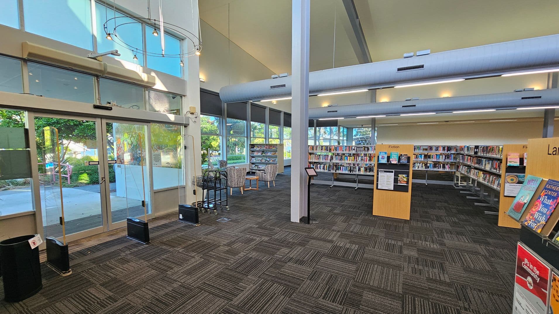 A library interior with large glass doors on the left, leading outside. The floor is covered with patterned carpet tiles in shades of grey and green. In the centre, there are bookshelves filled with books, labelled "Fiction." To the right, more bookshelves and a display stand with books. The ceiling is high with exposed ducts and modern lighting. Large windows on the far wall let in natural light, and there are two armchairs with a small table near the windows.