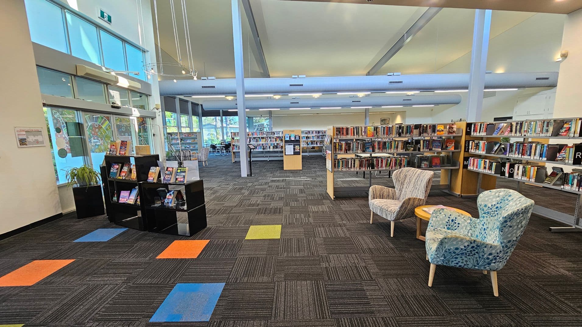 A library interior with a modern design. On the left, there are large windows with decorative patterns and a black display shelf holding books. The floor has a patterned carpet with colourful square accents in orange, blue, and green. In the centre, there are bookshelves filled with books. On the right, two patterned armchairs and a small round table are placed near more bookshelves. The ceiling is high with exposed beams and hanging lights.