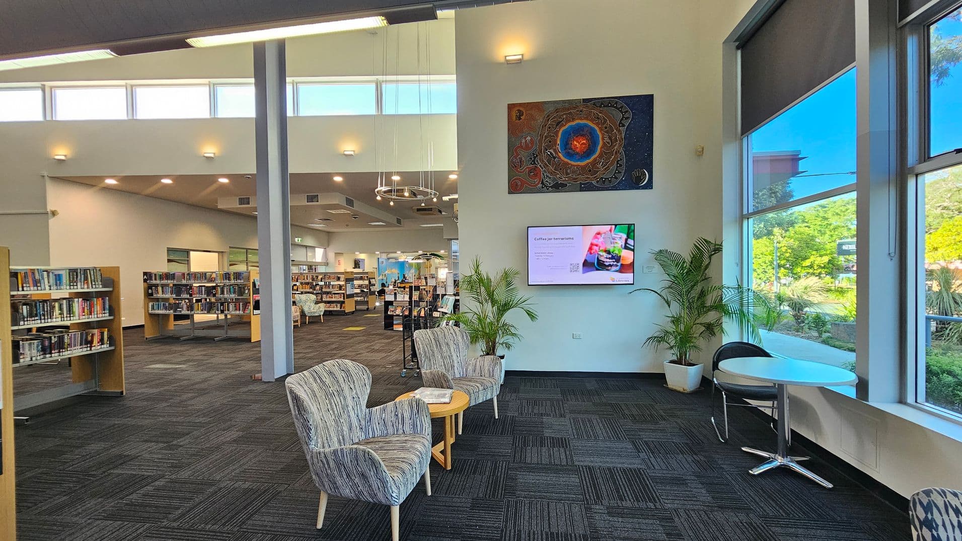 A modern library interior with a spacious layout. On the left, there are bookshelves filled with books. In the centre, two patterned armchairs and a small round table are placed near a wall with a digital screen and artwork above it. On the right, large windows allow natural light to illuminate the space, with a small round table and chair nearby. The floor is carpeted in a dark pattern, and the ceiling features recessed lighting and a circular chandelier.