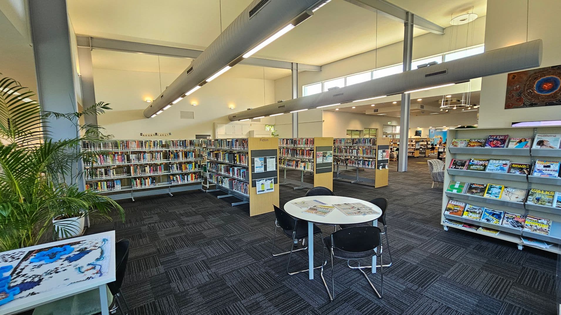 A spacious library interior with a high ceiling and large windows. On the left, there are tall bookshelves filled with books and a potted plant. In the centre, a round table with chairs has newspapers on it. On the right, a magazine rack displays various colourful magazines. The floor is carpeted with a dark pattern, and overhead, long light fixtures hang from the ceiling. In the background, more bookshelves and seating areas are visible.