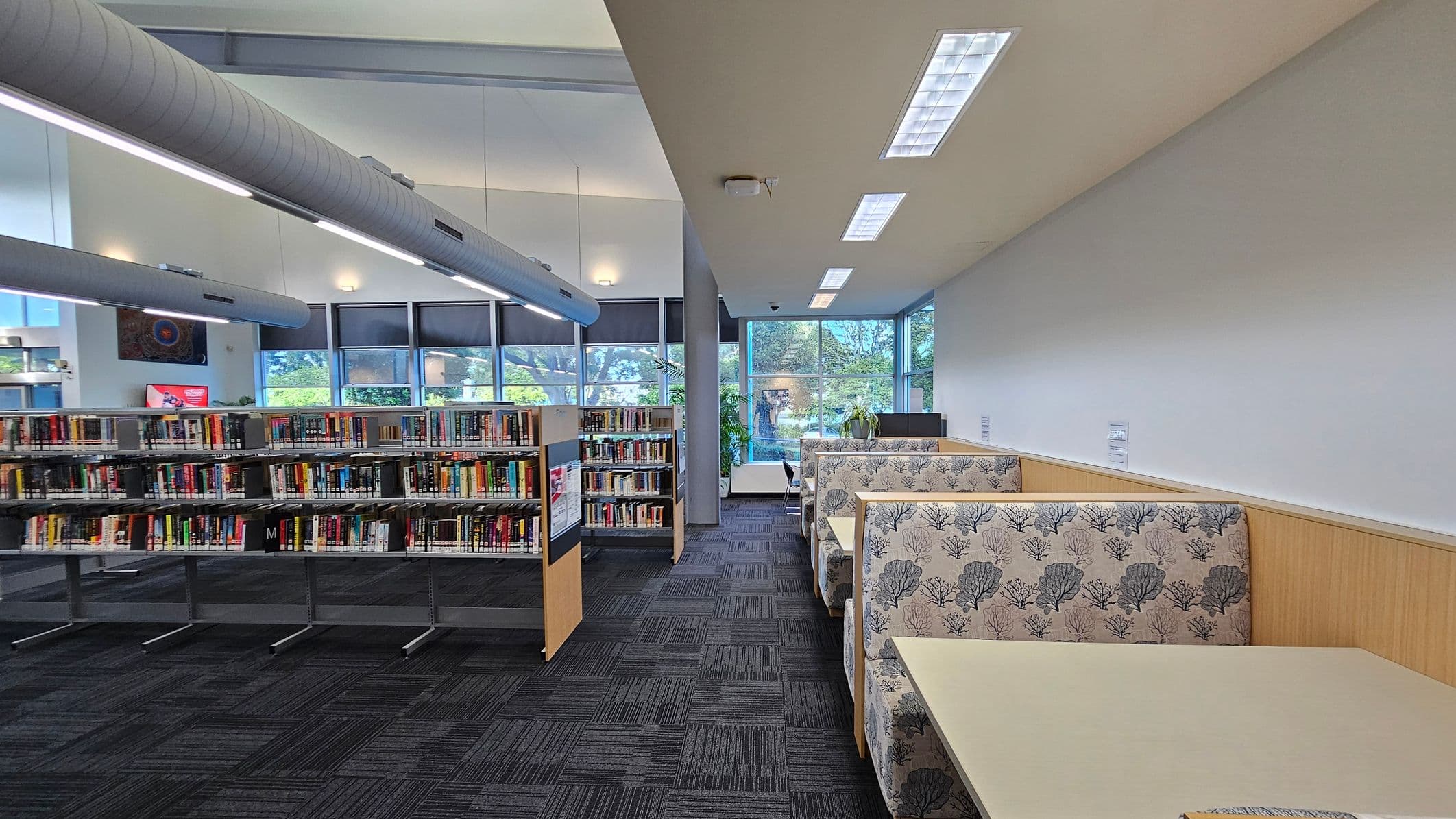 Library interior with bookshelves on the left filled with books, and a row of cushioned booths on the right with floral-patterned upholstery. The floor is carpeted in a dark, patterned design. Large windows in the background allow natural light to enter, with views of trees outside. The ceiling features long, cylindrical air ducts and recessed lighting.