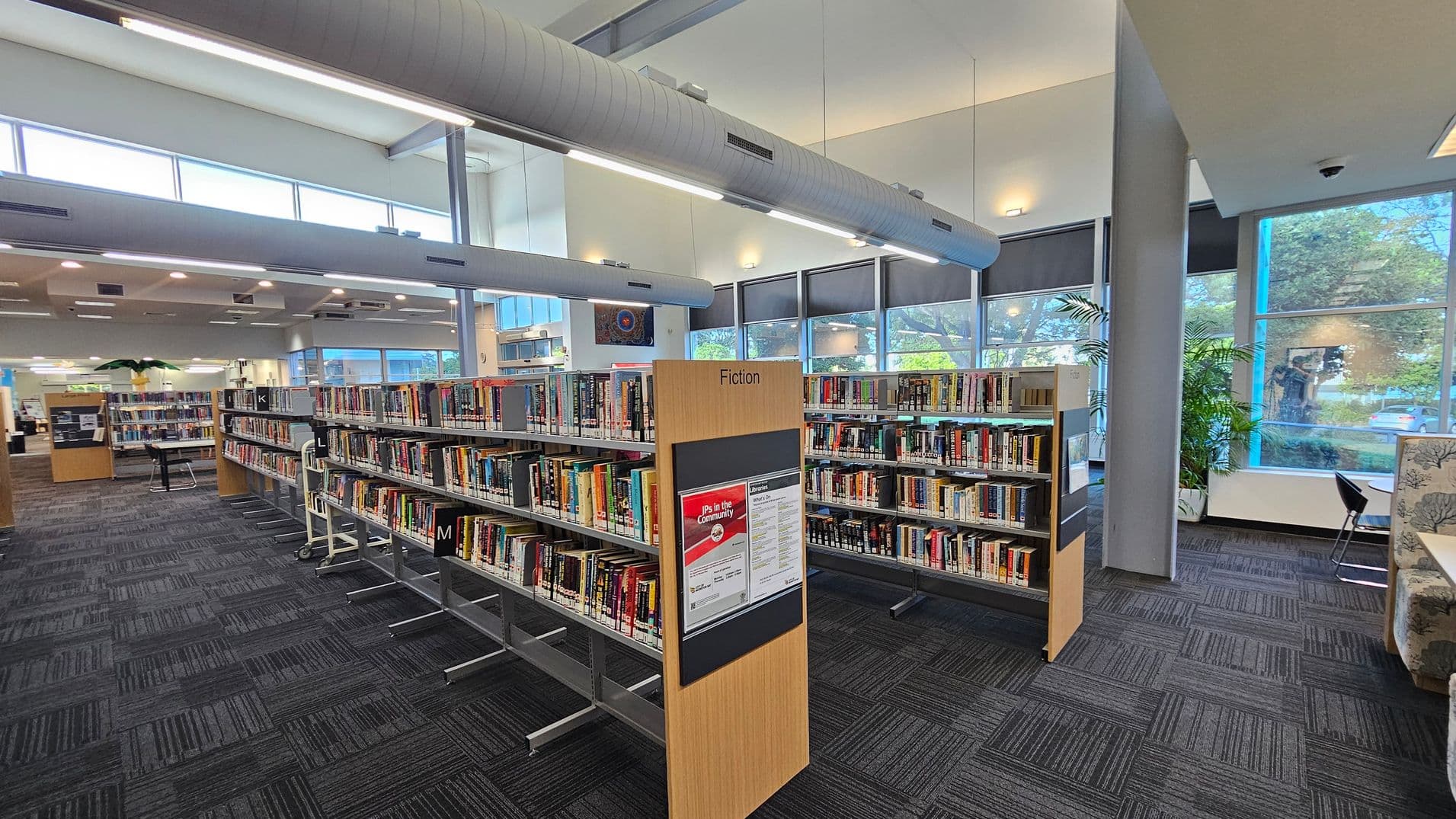 Bookshelves filled with books are arranged in rows on a carpeted floor, with a sign labelled "Fiction" in the foreground. Large windows on the right allow natural light to illuminate the space, and there are potted plants near the windows. The ceiling features exposed ductwork and fluorescent lighting. In the background, more bookshelves and a seating area with chairs and tables are visible.