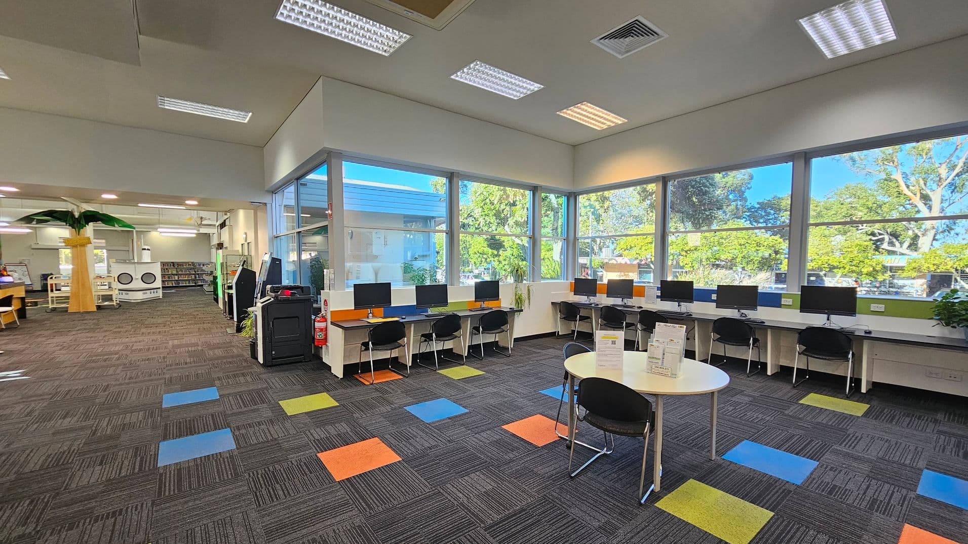 A modern library interior with large windows on the right, allowing natural light to illuminate the space. Several computer workstations with black chairs are lined up against the windows. In the foreground, a round table with informational pamphlets and chairs is visible. The floor is carpeted with a pattern of grey tiles interspersed with colourful squares. On the left, there is a tall artificial palm tree and bookshelves. The ceiling has recessed lighting and air vents.