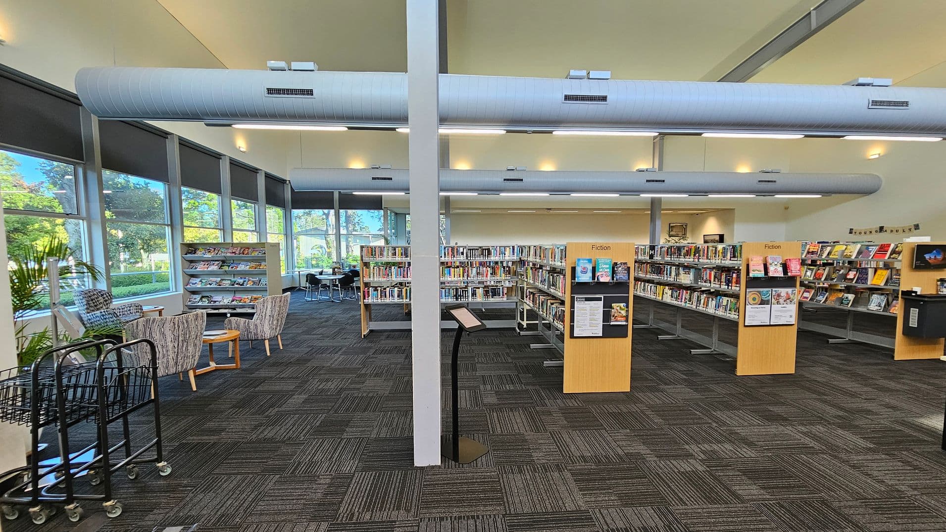 A library interior with a grey and green patterned carpet. On the left, there are large windows with roller blinds, a magazine rack, and seating with patterned chairs and a small table. In the centre, a vertical support beam stands in front of a digital kiosk. Bookshelves labelled "Fiction" are filled with books. On the right, more bookshelves and a section labelled "Young Adult" are visible. The ceiling features large cylindrical air ducts and recessed lighting.