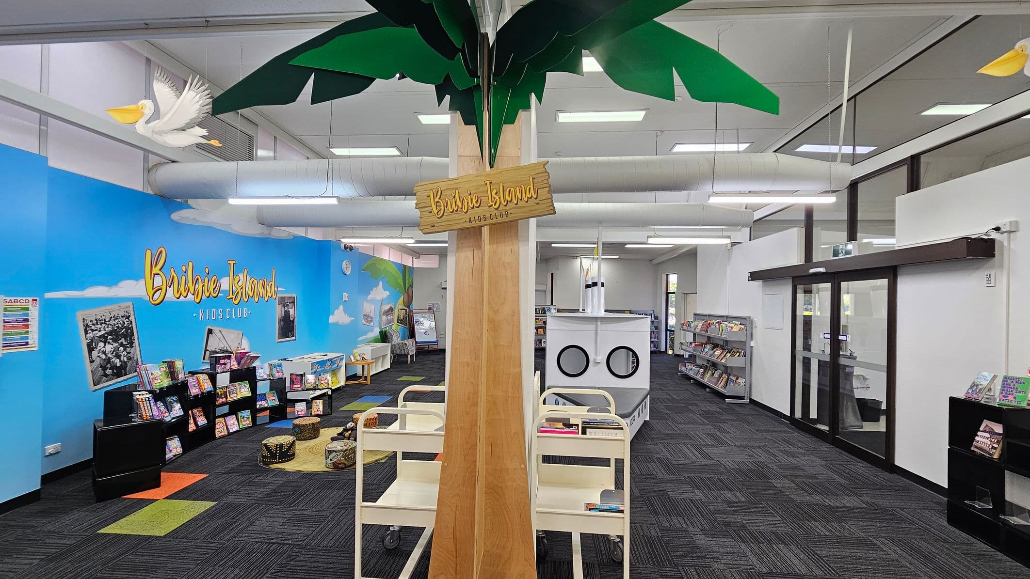 A library space with a central wooden column featuring a "Bribie Island Kids Club" sign. Above, green leaves and a pelican decoration are visible. On the left, a blue wall displays the same "Bribie Island" text, with books on black shelves and colourful seating on a patterned carpet. On the right, glass doors and more bookshelves are present. The ceiling has exposed ducts and fluorescent lights, and the floor is covered with dark carpet tiles.