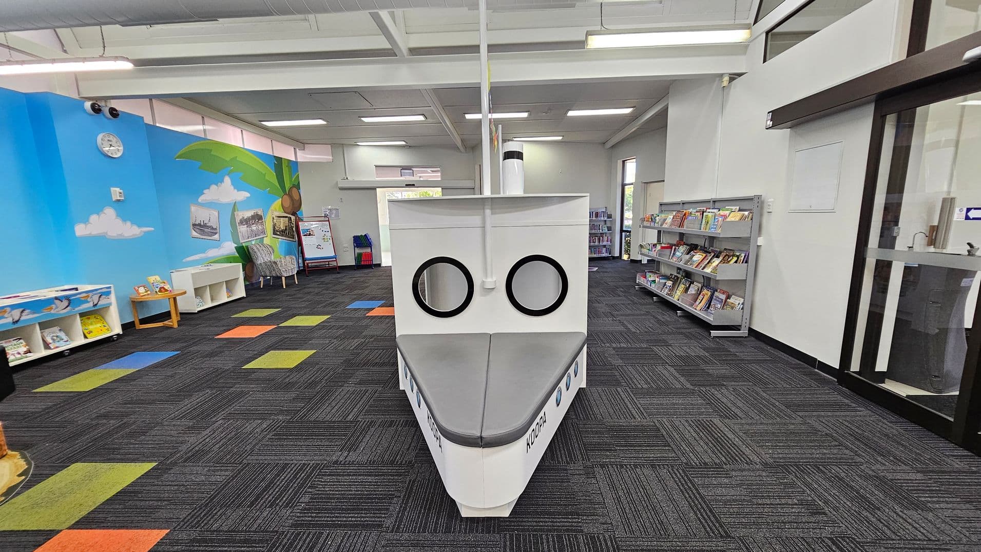 A library space with a boat-shaped reading nook in the centre, featuring two circular windows and grey seating. The left wall is painted with a blue sky, clouds, and a palm tree, with bookshelves and a clock. The right side has bookshelves filled with books and a glass door. The floor is carpeted in dark grey with colourful square patterns, and the ceiling has white panels with fluorescent lights.